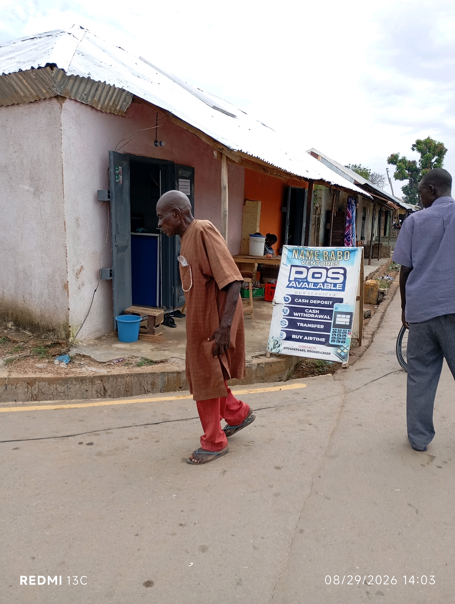 In Shendam, Nigeria, an elderly man wearing a brown robe and red pants walks on a paved road beside a building with a corrugated metal roof. A white A-frame sign advertising "NAME RABO VENTURES POS AVAILABLE" and listing services like "CASH DEPOSIT," "CASH WITHDRAWAL," "TRANSFER," and "BUY AIRTIME" stands on the sidewalk. Another man in a blue shirt and grey pants walks away from the camera on the same road. The environment appears to be a commercial area with multiple businesses lining the street.
