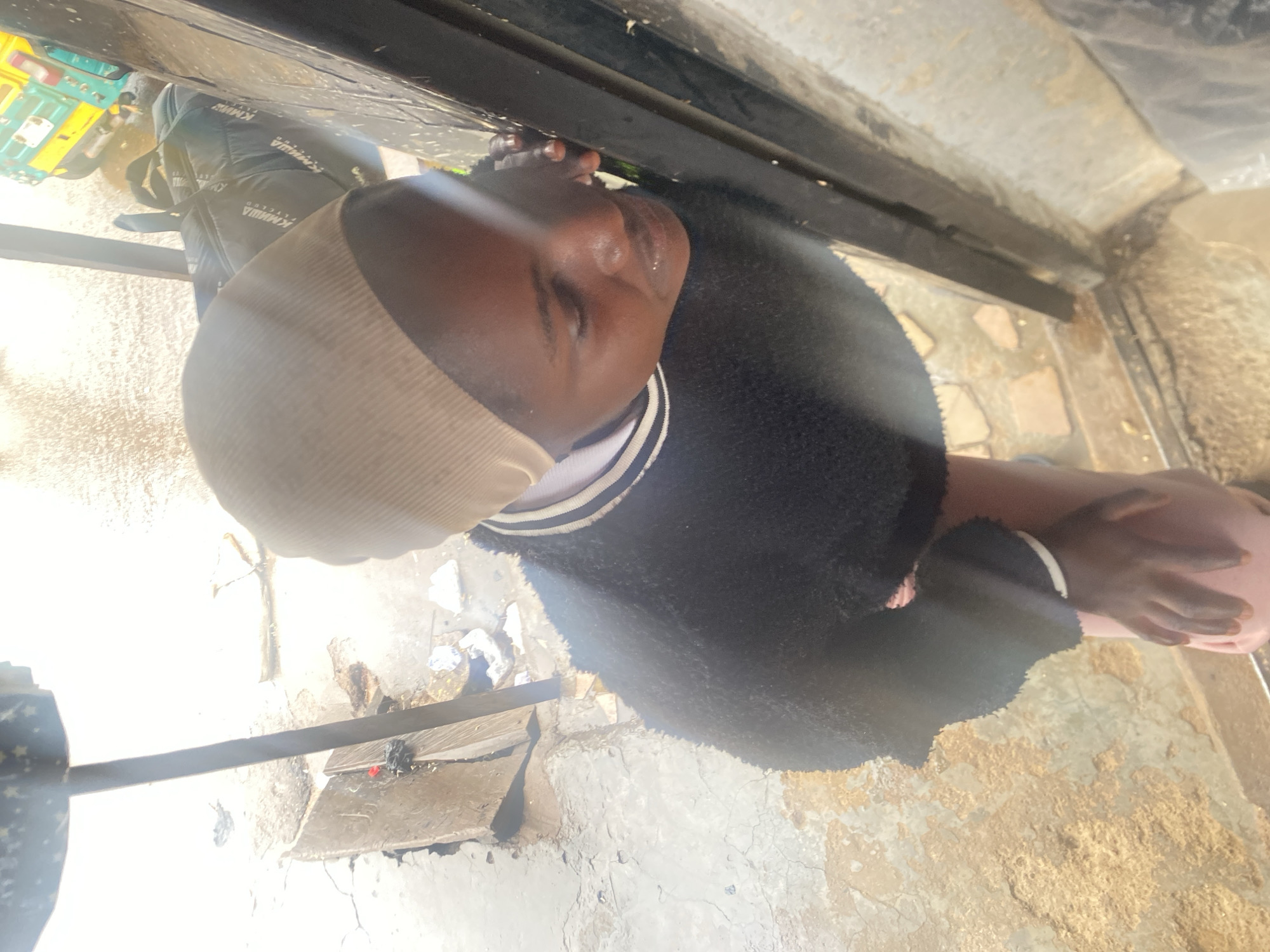 A person is shown from an overhead perspective. The individual is wearing a light-colored head covering, a striped shirt or collar, and a black fuzzy jacket. The surroundings appear to be a rough concrete or stone surface with debris scattered on it. Part of a vehicle or structure is visible at the top of the frame. The location is within the city of Bukuru, Nigeria.
