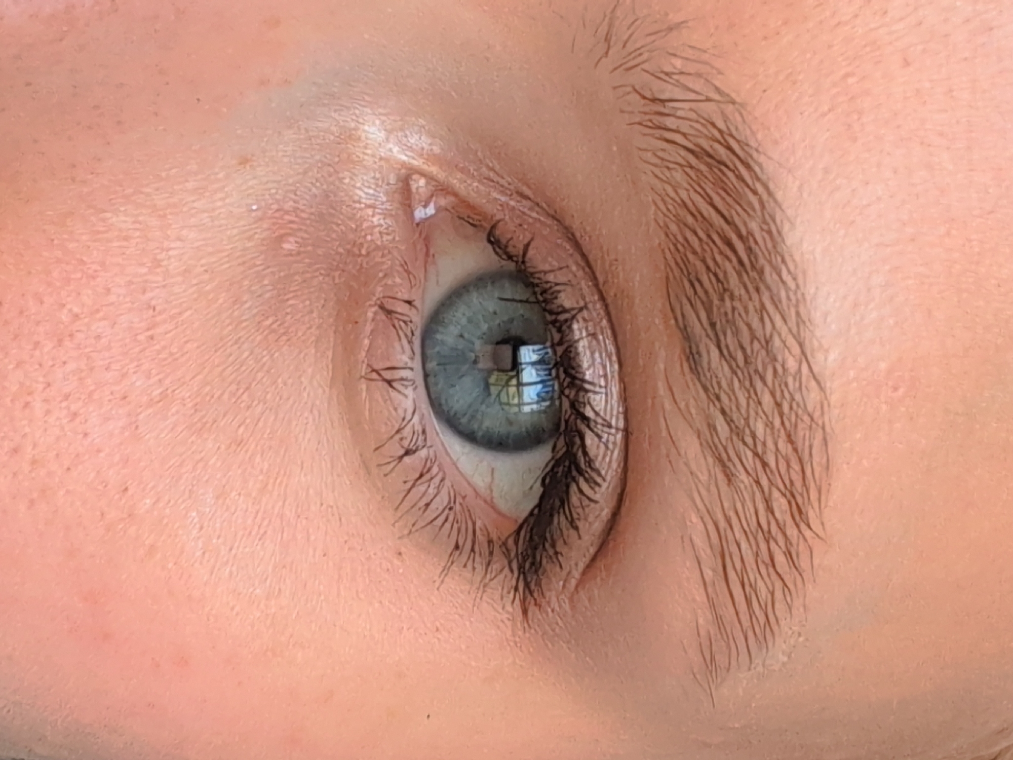 The image is a close-up shot of a person's eye. The iris is a pale blue-green with flecks of grey and a dark pupil. The surrounding skin is fair with visible pores and some light blemishes. The eyelashes are dark and appear to have mascara applied. The eyebrow is light brown and somewhat sparse. The background is out of focus and appears to be skin. There is a reflection in the pupil of the eye, possibly from a window or light fixture. The location in Bugry, Russia is not discernible from the image itself.