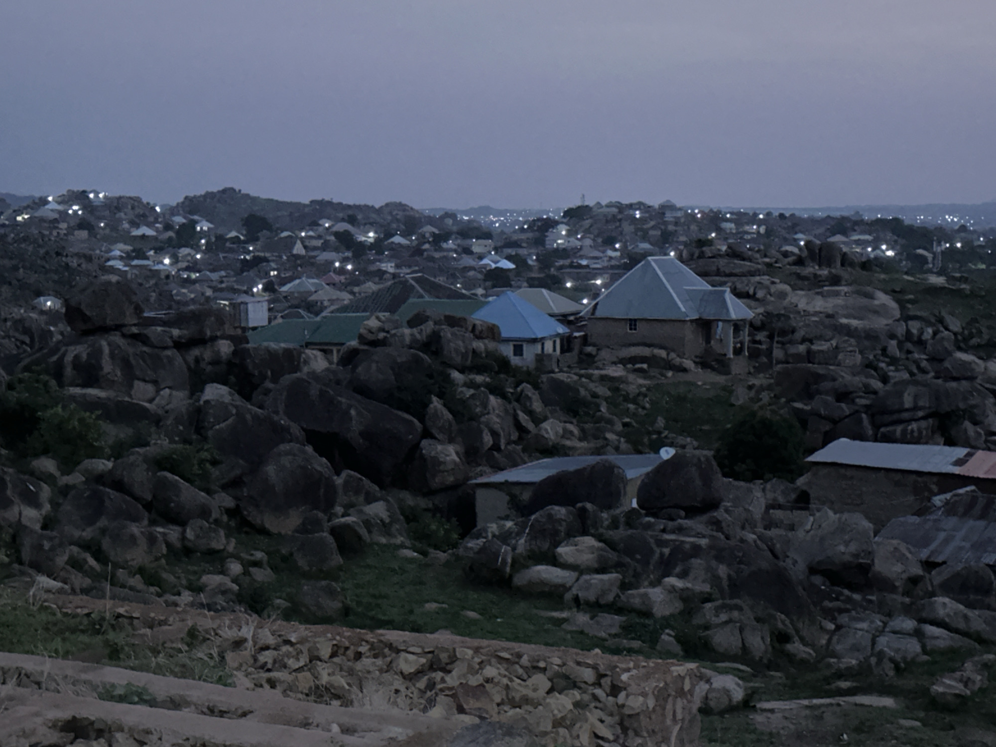The image shows a section of Jos, Nigeria at dusk, marked by a rocky landscape interspersed with modest residential buildings. The foreground consists of numerous dark, irregularly shaped rocks and sparse vegetation. In the midground, several houses with varying roof colors (blue, green, grey) are visible, built among the rocks. The background features a dense collection of buildings, many with lights on, stretching into the distance. The sky is a uniform grey, indicating early evening.