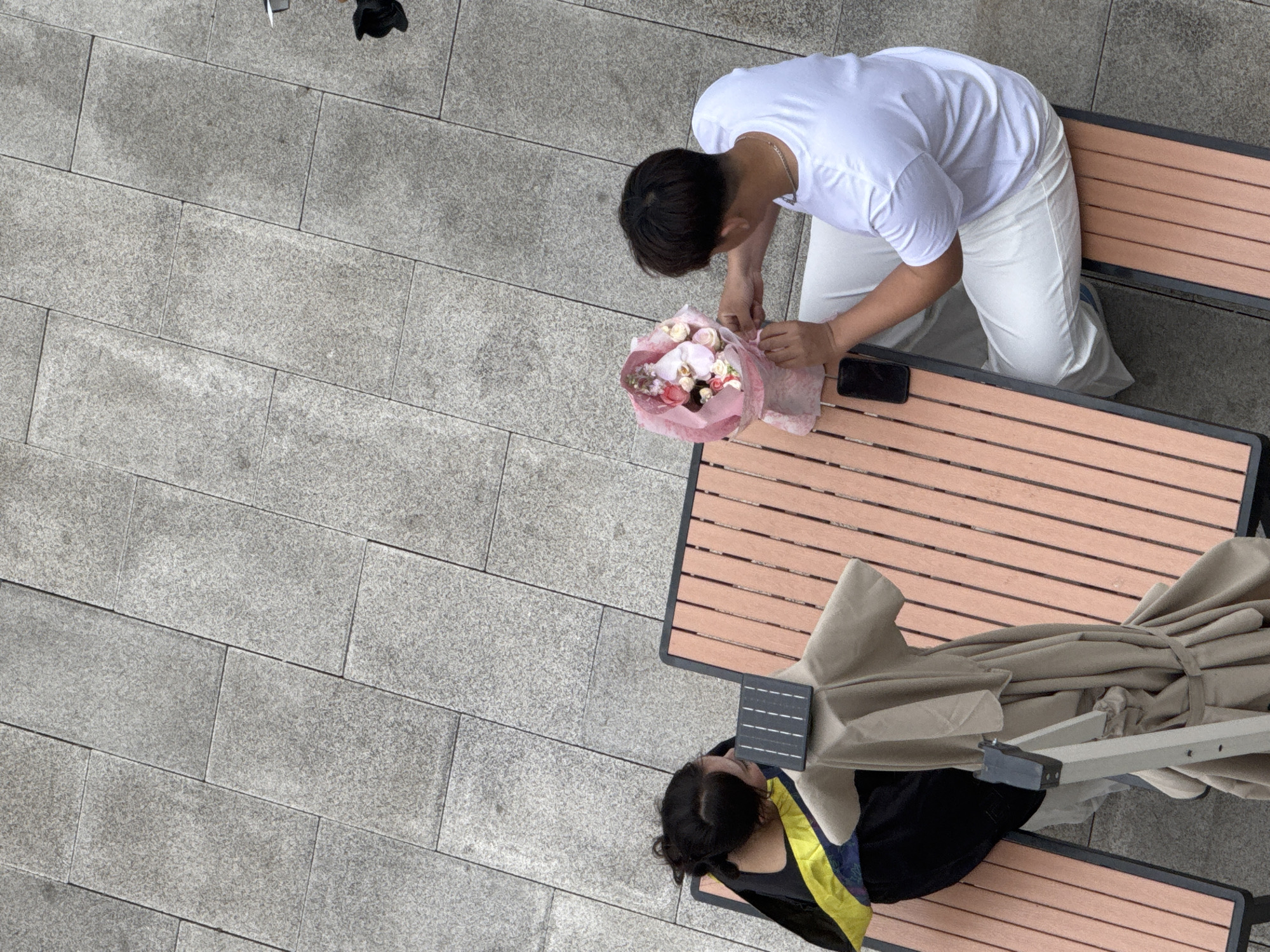 An overhead shot shows two people sitting on park benches on gray paved ground in Zhongtun, China. One person, facing the right side of the image, is wearing a white t-shirt and white pants. They are arranging a bouquet of flowers on a wooden table placed in front of them. A black cell phone sits on the table next to the flowers. Another person is sitting on a bench on the bottom right of the image, wearing a black top with a yellow design. An umbrella, wrapped in a beige cloth, is positioned next to them.