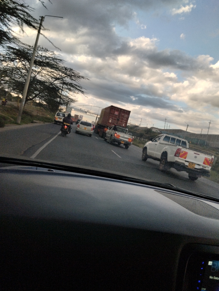 The image shows a view from the driver's seat of a car traveling on a four-lane highway in Mlolongo, Kenya, located at coordinates -1.407 latitude and 36.947 longitude. The sky is overcast with white clouds. The road is paved and has a white dividing line. There are five vehicles in the scene: a white truck, a white pickup truck, a silver car, a red car, and a motorcycle. The white truck is traveling in the left lane, the pickup truck is in the right lane, and the motorcycle is in the left lane. The silver car and the red car are both in the right lane. The motorcycle is driven by a person wearing a helmet. There are no other people visible in the image. The environment is suburban, with tall grass on either side of the road. There are power lines running along the side of the road. The vehicles are moving in the same direction as the camera. There is no interaction between the vehicles or the people. The image appears to have been taken at midday, with a slight haze.