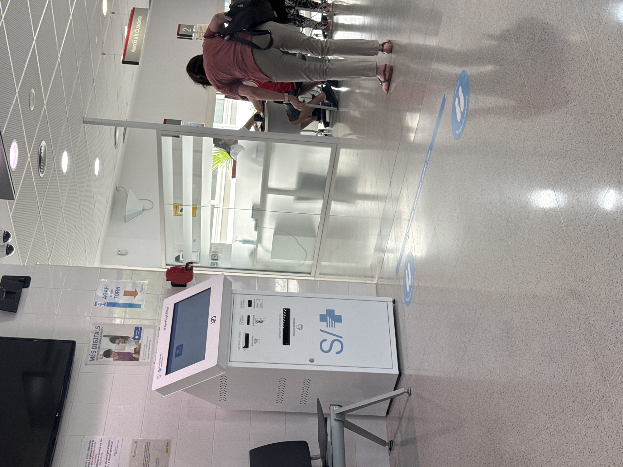 The image depicts the interior of what appears to be a public service office or healthcare facility. In the foreground, a white self-service kiosk with a touchscreen display is positioned to the left. The kiosk has various labels and symbols, including "S/+" and "Institut Català de la Salut." To the left of the kiosk is a grey metal chair with a black seat. The floor is a speckled grey material with a tiled pattern, and blue social distancing markers are visible on the floor, including a circular decal with two footprint icons.
In the middle ground, a glass partition separates the area with the kiosk from another section of the building. Through the glass, a desk is visible with individuals behind it, suggesting a service counter. To the right, a woman wearing a pink t-shirt and beige pants is bent over, interacting with something on the floor. She has a dark bag slung over her shoulder. Further to the right, other individuals are partially visible. A sign with the text "Atenció a la Ciutadania" (Citizen Attention) is mounted on the wall in the background. The environment appears to be well-lit with artificial lighting. The location is identified as L'Hospitalet de Llobregat, Spain.