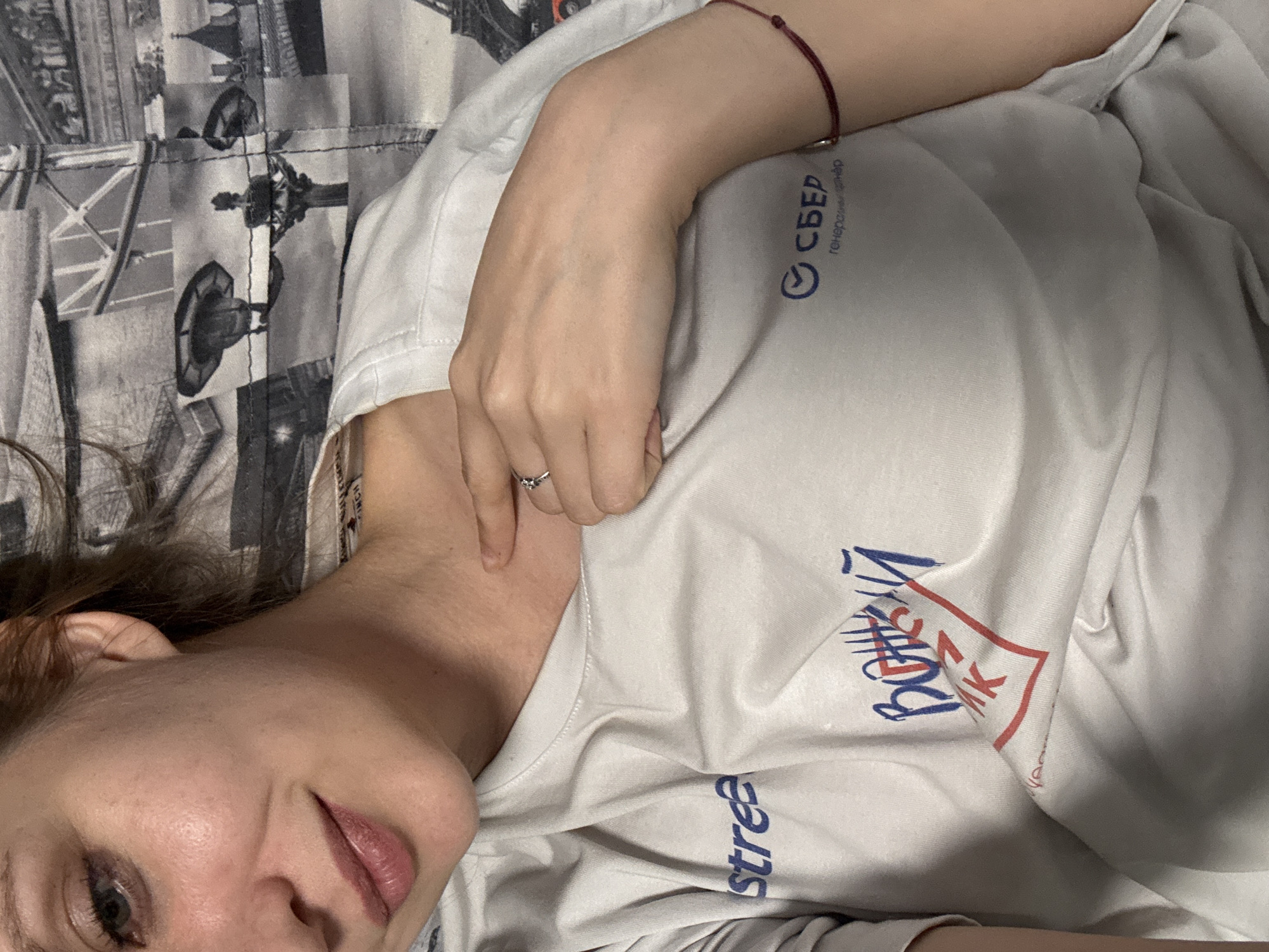 A close-up, medium shot shows a young Caucasian woman lying down and looking up towards the camera. She is wearing a white t-shirt with blue and red lettering and graphics. The t-shirt has a dark maroon bracelet on her right wrist. Her left hand is resting on her chest. The background is a blurred pattern of black and white images of buildings and structures. The lighting is soft and natural. The overall impression is casual and relaxed.