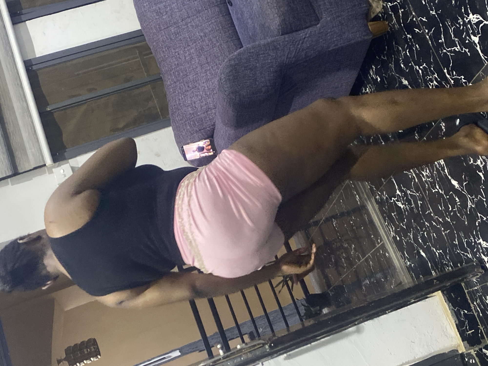 An overhead, tilted shot captures a person lounging indoors, likely in a residence in Abuja, Nigeria, during the evening or night. The main subject is a person, appearing to be female, lying on their side with their back mostly to the viewer, wearing a black sleeveless top and pink shorts. Their dark skin tone is visible on their arms and legs.
A dark purple, textured armchair or sofa occupies a significant portion of the frame. A smartphone, displaying an indistinct image or video, rests on the sofa near the person's hip. The flooring is distinctive, featuring black tiles with white, marble-like veins.
In the background, a window with horizontal dark frames and blinds is visible on a white wall, next to what appears to be a light switch. A metal railing is present in the middle ground, suggesting the person might be on a balcony or a raised indoor area overlooking another space. Beyond the railing, the background is dark and blurry, possibly showing an outdoor night scene or a dimly lit interior. The overall lighting is subdued, indicating a lack of bright daylight. No discernible text is visible beyond the indistinct content on the smartphone screen. The person's face and emotions are not visible.