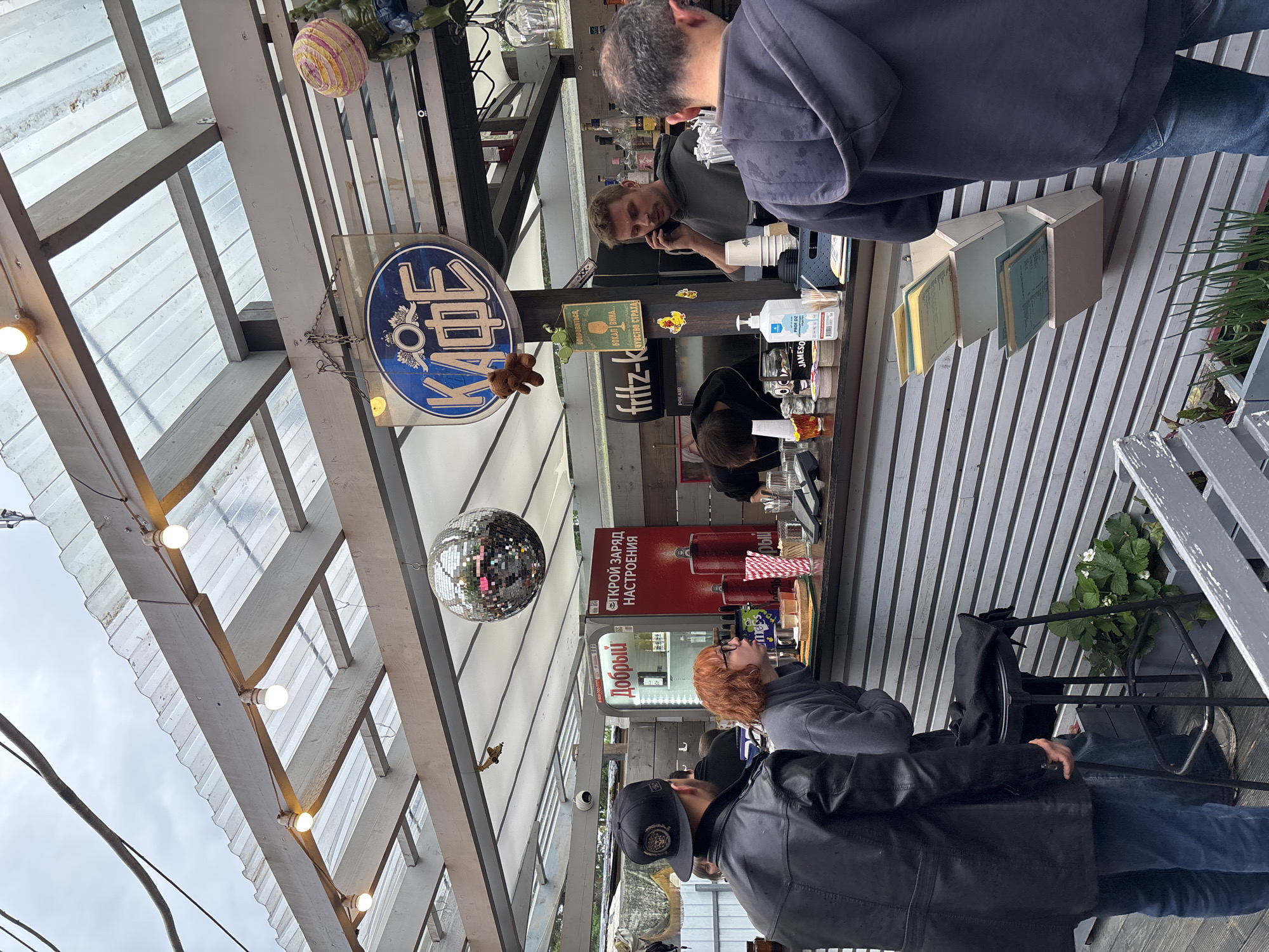 The image shows an outdoor cafe with a covered seating area. There are several people at the cafe. One man is standing behind the counter, talking on his phone. Another man is sitting at the counter, looking at his phone. A woman with red hair is standing at the counter, and a man in a black leather jacket is standing next to her. The cafe has a disco ball hanging from the ceiling, and there are bottles of alcohol behind the counter. The cafe appears to be located in the city of Komarovo, Russia, as indicated by the sign that says "КАФЕ" (Cafe).