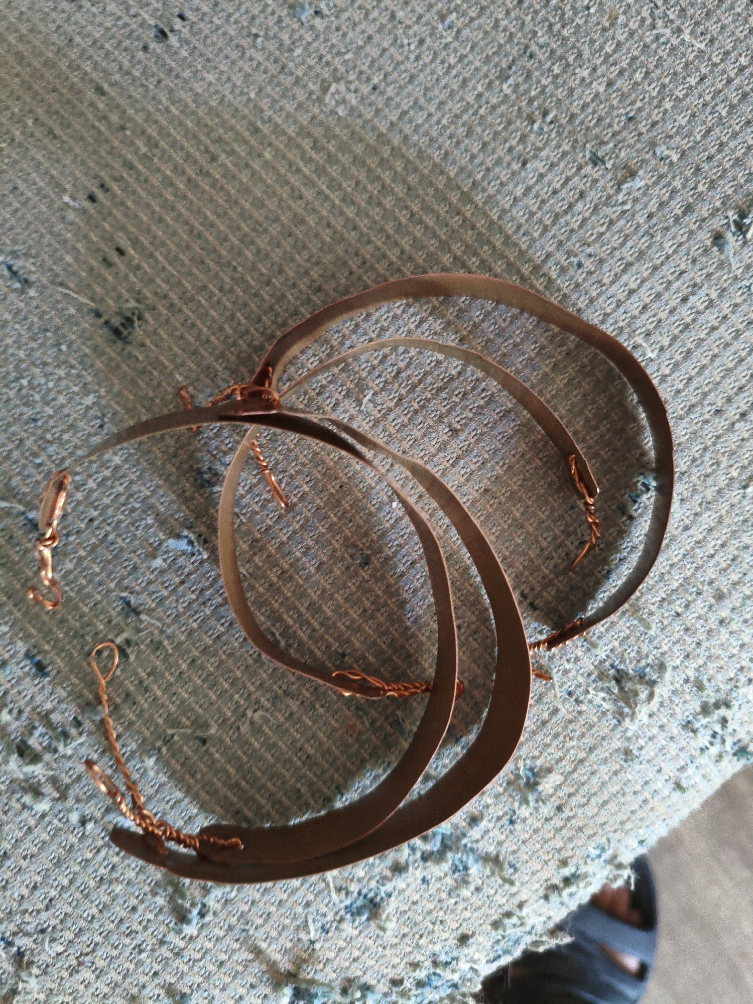 Three curved, copper-colored metal strips are arranged in a circular, overlapping pattern on a textured, light blue fabric surface. The metal strips are thin and appear to be made of a malleable material, possibly for crafting or jewelry. One of the strips has a small copper-colored hook attached to its end. The fabric surface has a rough texture with visible threads and small dark specks. In the lower right corner of the image, a portion of a person's foot wearing dark blue sandals is visible, suggesting the scene is indoors or in a shaded outdoor area. The overall lighting is soft, casting subtle shadows. There are no discernible landmarks or identifying features that would indicate a specific location in Dessau, Germany.