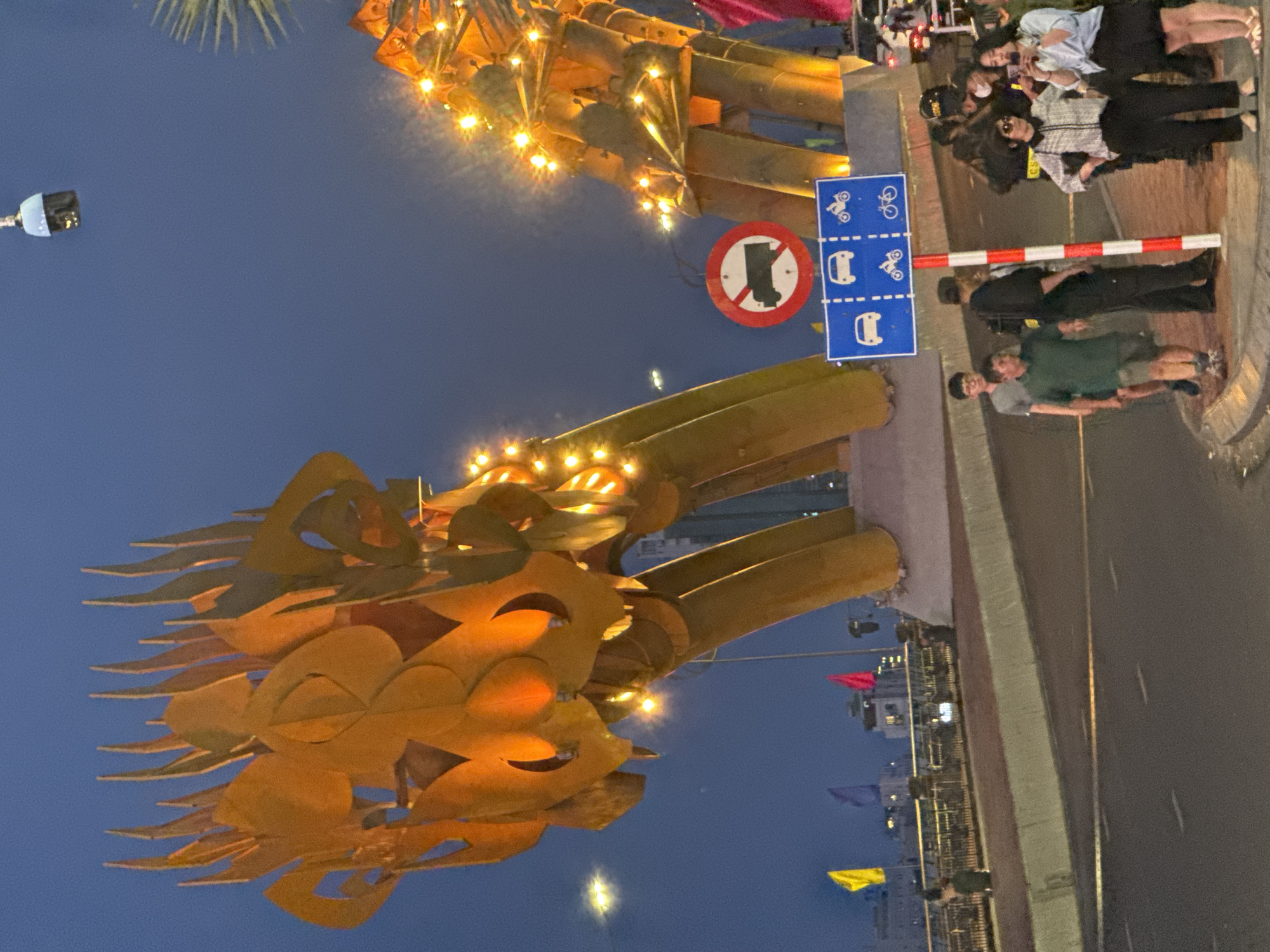 The image depicts the illuminated head of the Dragon Bridge in Đà Nẵng, Vietnam, at night. The dragon head is gold in color and features intricate metalwork. To the right, on the bridge's walkway, a group of people are standing, some of whom appear to be taking photos or interacting with each other. Near them, street signs indicate regulations about vehicles and bicycles. The road is visible alongside the walkway, and in the background, buildings and flags can be seen along the riverfront. The sky is a dark blue, suggesting it is either dusk or night.