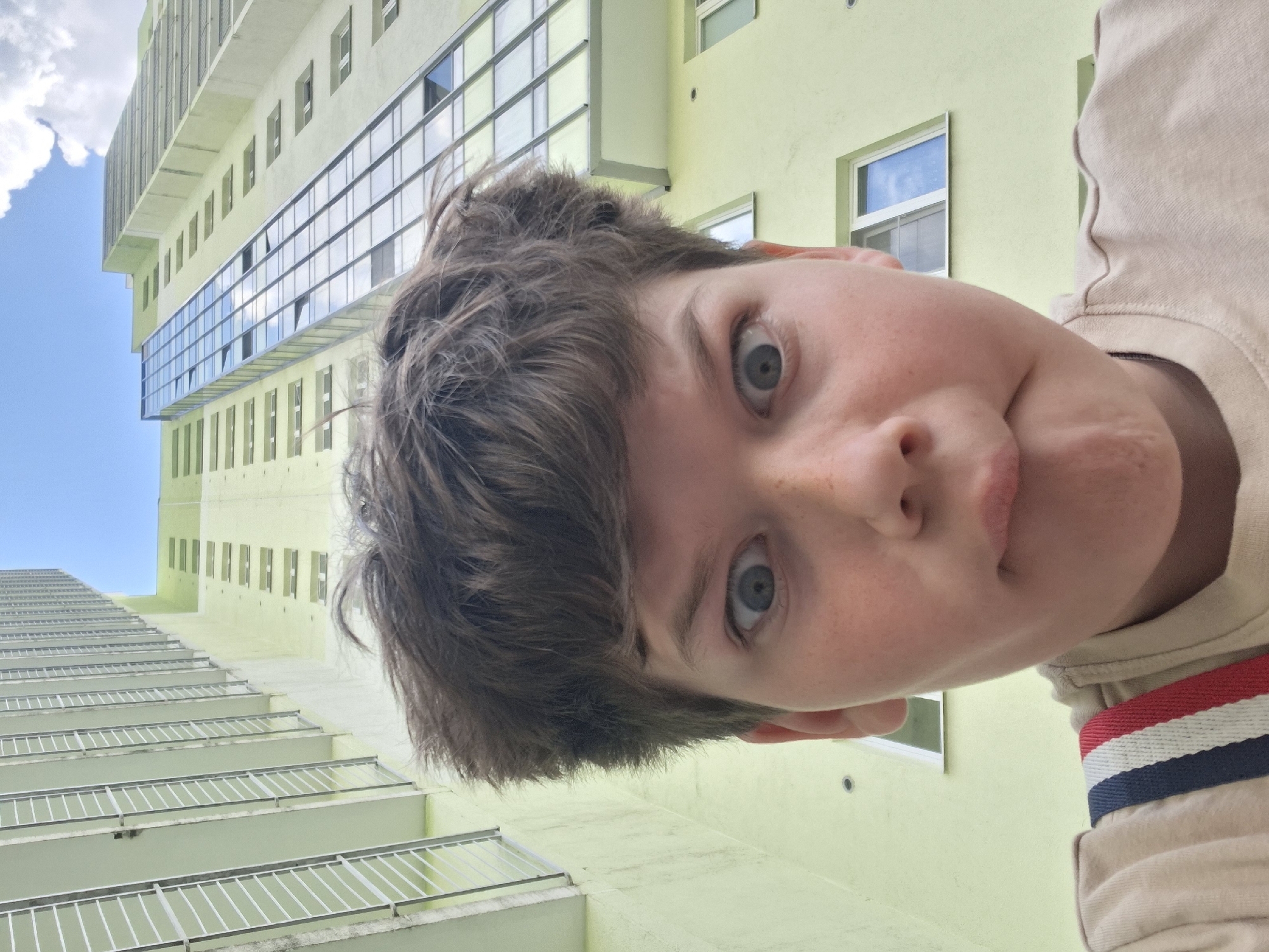 This image, taken from a low, upward-looking perspective, features a person's face prominently in the foreground with a tall, modern building rising dramatically into the blue sky behind them.
The primary subject is a young person, likely a child or teenager, whose face occupies the right half of the frame. They have light skin, freckles, and light-colored eyes, looking directly at the camera with a wide-eyed, somewhat surprised or playful expression, and a slightly puckered mouth. Their brown hair is tousled, framing their forehead. They are wearing a light-colored (possibly beige or cream) long-sleeved top, with a distinctive red, white, and blue striped band visible on the sleeve near their shoulder.
The setting is an urban environment, specifically against a tall, multi-story building that appears to be an apartment or office block. The building is painted a uniform, pale lime green color. It features numerous rows of windows, some of which are large and reflective, showcasing reflections of the sky and clouds. On the lower section of the building, there are multiple balconies with thin, white or metallic railings extending outwards, creating a patterned effect. The style of the architecture suggests a modern structure, possibly found in a city like Saint Petersburg, Russia, given the provided context.
The activity appears to be a casual selfie or a portrait taken by someone close to the subject. The person's direct gaze and playful expression suggest a candid and informal moment.
Notable details include the bright daylight and clear blue sky with fluffy white clouds, indicating good weather. The low-angle shot emphasizes the height of the building and creates a sense of depth and scale, with the person's face appearing large in comparison to the background. There is no visible text in the image.