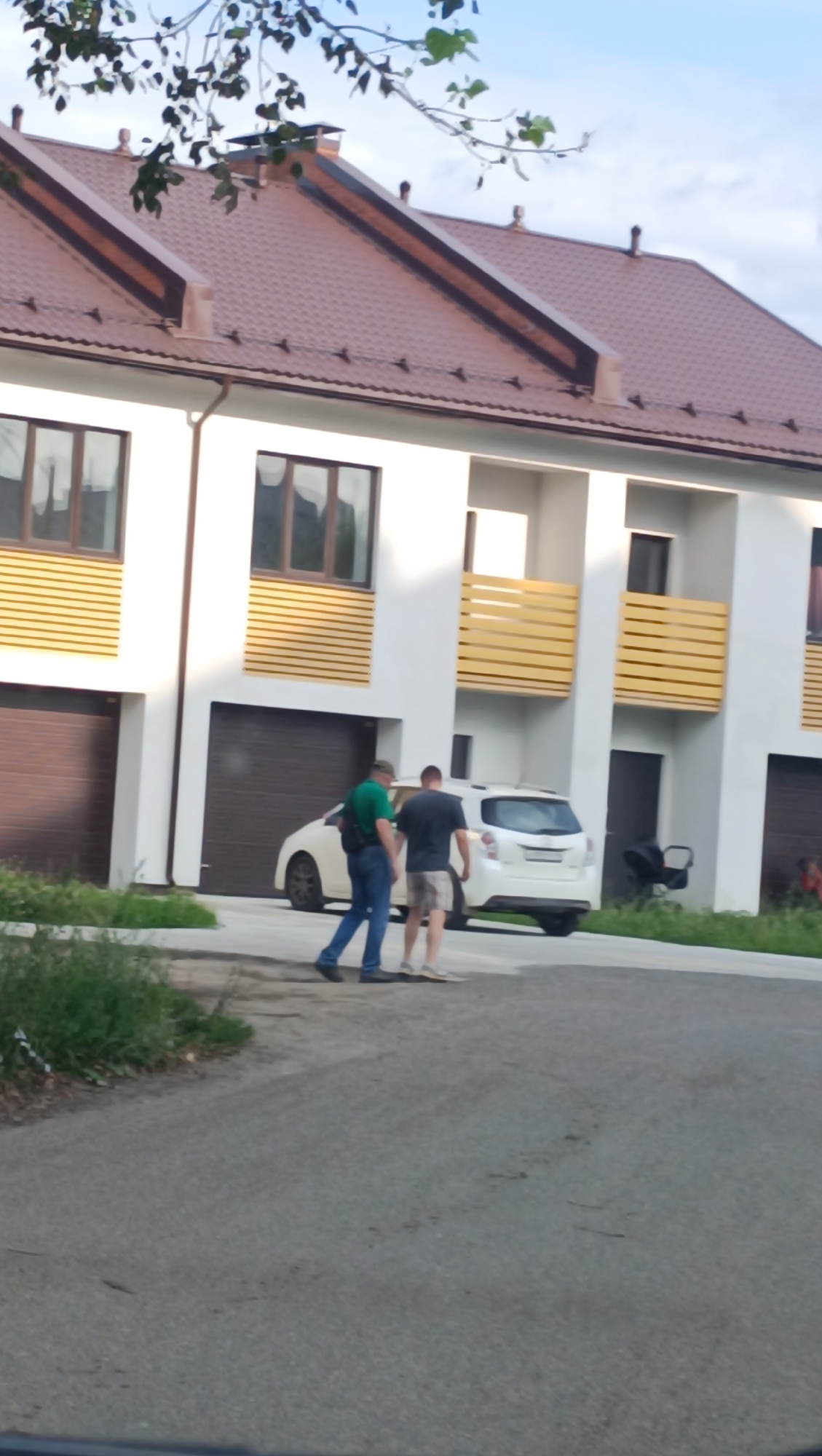Two males are walking on an asphalt road in front of a row of modern townhouses with brown roofs. The townhouse on the left has a brown garage door and a yellow-slatted balcony on the second floor. The townhouse in the center has a white SUV parked in front of its brown garage door, and a yellow-slatted balcony on the second floor. The townhouse on the right has a black stroller parked outside and a yellow-slatted balcony on the second floor. The sky is partly cloudy and blue. The presence of these townhouses suggests this is a residential area, possibly a newer development. The foliage in the foreground indicates a green, somewhat unkempt area adjacent to the road. This scene is consistent with a residential street in Chaikovsky, Russia, likely an urban or suburban setting given the modern construction.