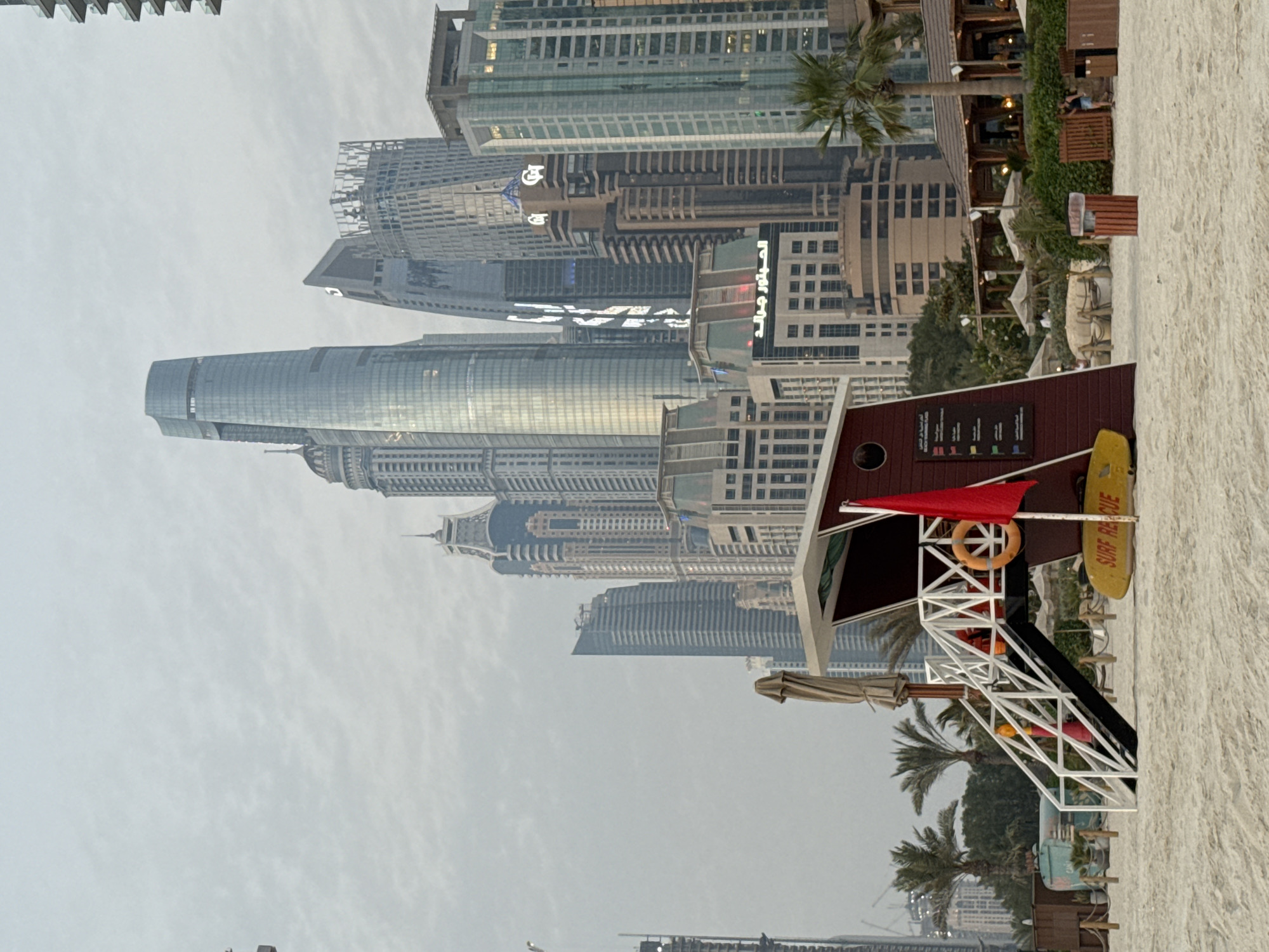 This image captures a view of the Dubai Marina coastline, United Arab Emirates. In the foreground, a red and white lifeguard tower stands on a sandy beach. A yellow surfboard with the words "SURF RESCUE" is leaning against the tower, and a red flag is flying from a pole. The background features a dense cluster of modern skyscrapers, characteristic of Dubai's urban landscape, under an overcast sky. Palm trees and beach umbrellas are visible around the lifeguard tower, suggesting a recreational area. A few people are visible in the distance, near some buildings and a beachside establishment.