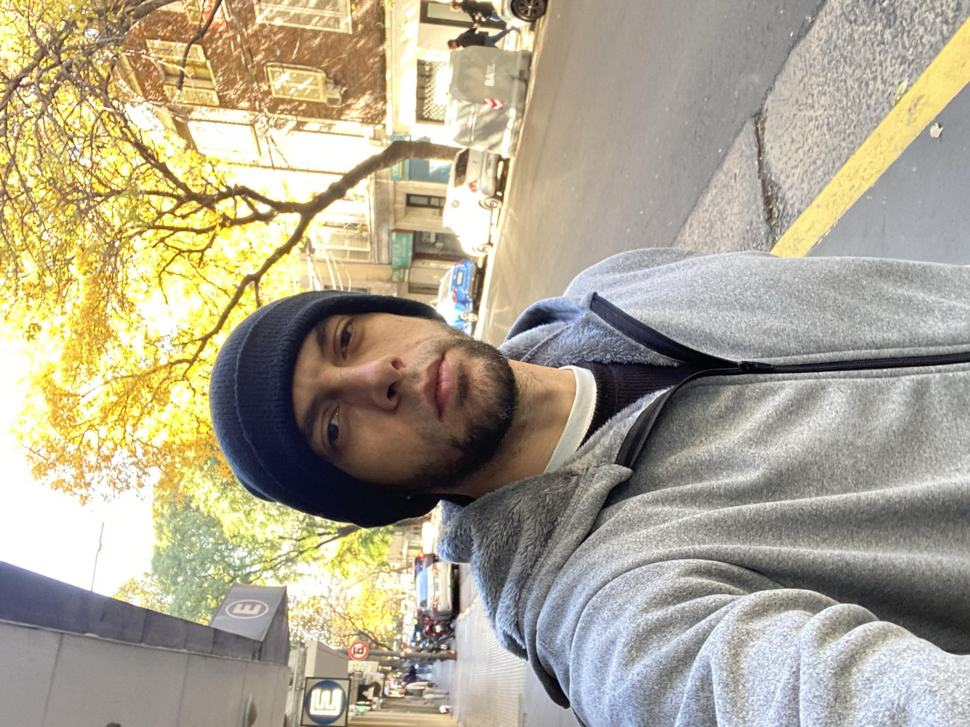 A man in a black beanie and gray fleece jacket is taking a selfie on a sidewalk in Buenos Aires, Argentina. The background includes a street with parked cars, pedestrians, buildings, and trees with yellowing leaves. There is a blue and white metro sign visible.