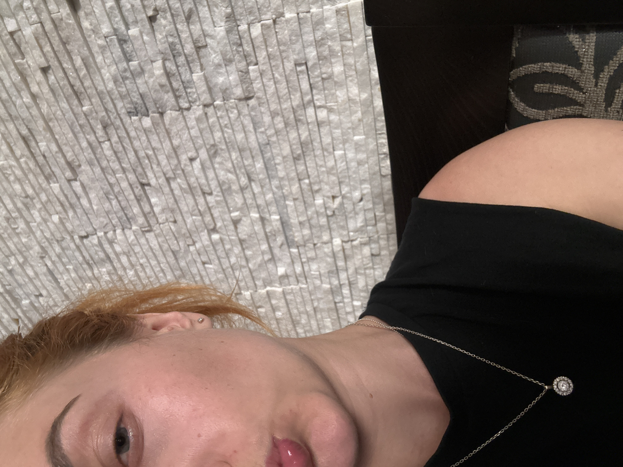 A close-up, tilted portrait features an individual, likely female, in an indoor setting in Budva, Montenegro. The person's head, right shoulder, and upper torso are visible, positioned against a light-colored, textured stone wall. They have reddish-blonde hair and a dark, shaped eyebrow. One eye is partially visible, and the lips are slightly parted, glossy, and pink. A small silver stud earring is visible in their ear.
The person is wearing a black garment, possibly a t-shirt or top, revealing a bare shoulder. A delicate silver chain necklace with a circular pendant, adorned with a central stone surrounded by smaller stones, rests on their chest. The background prominently features a wall composed of light grey or off-white rectangular, rough-cut stone tiles arranged in a vertical stacked pattern, providing a rugged texture. To the right, a dark object, possibly part of a chair or sofa with a subtle dark grey patterned upholstery, is visible. The lighting is soft and even, suggesting an indoor environment. The person's expression appears neutral or contemplative, and no specific activity is depicted beyond being photographed. No visible text is present in the image.