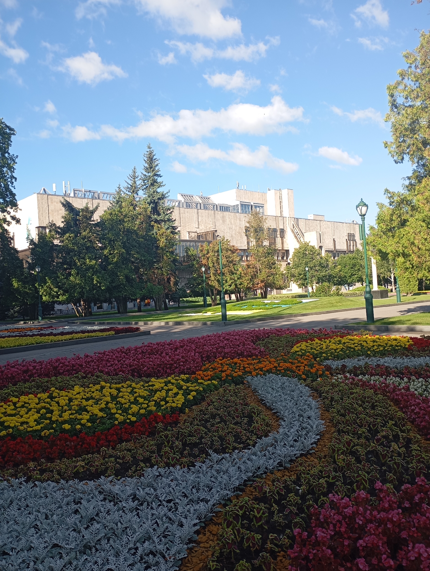The image shows a large, modern building with a textured facade, surrounded by trees and a meticulously arranged flower garden in the foreground. The building appears to be an institutional or public building, possibly a university or government building. The flower garden features several rows of colorful flowers, including red, yellow, orange, pink, and white varieties, arranged in a curved pattern. The sky is blue with scattered white clouds. The presence of mature trees suggests a well-established park or public space. The overall scene is tranquil and well-maintained. Based on the architectural style and landscaping, this location is likely in the city of Kharkiv, Ukraine, possibly on the grounds of Kharkiv National University or a similar institution known for its extensive green spaces and public monuments.