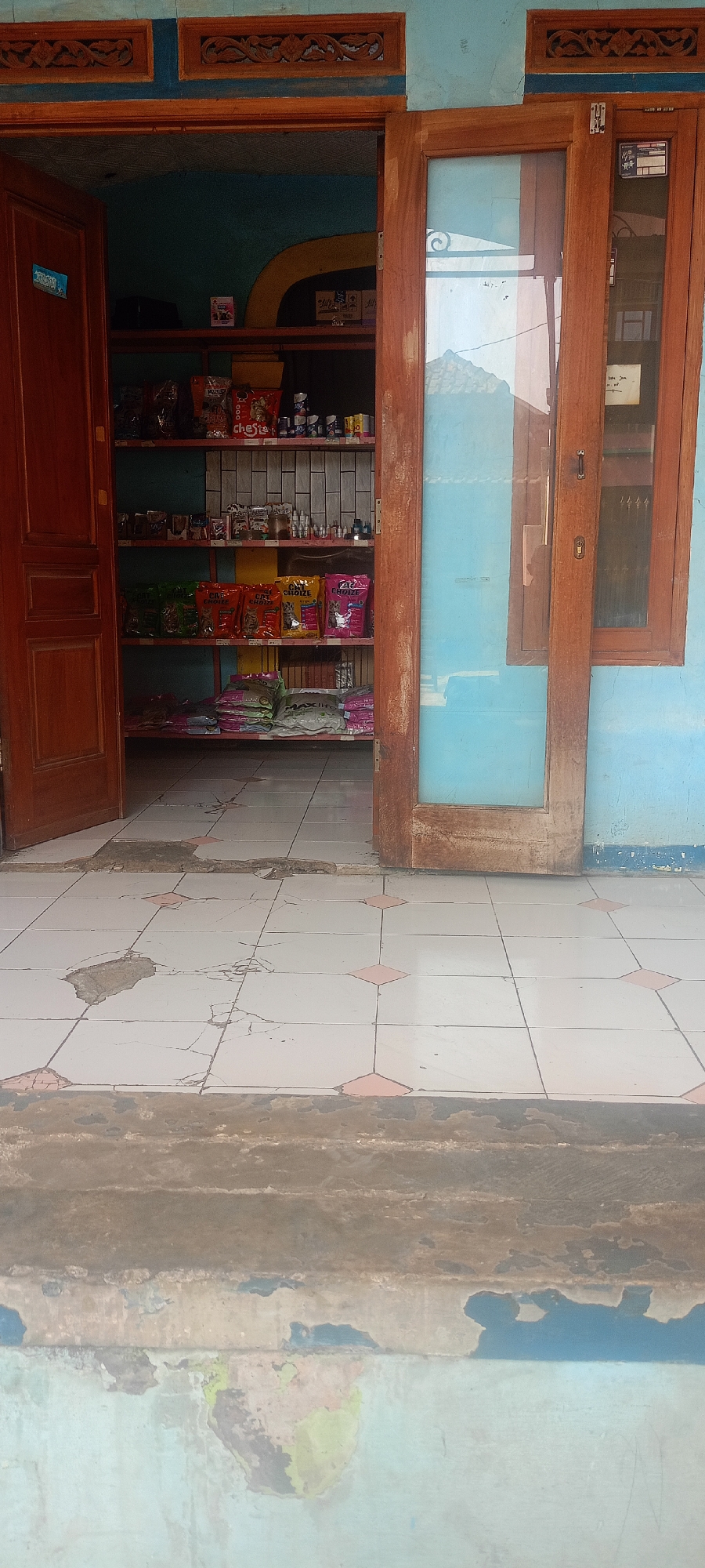 The image shows the exterior entrance to a small shop, likely in Padalarang, Indonesia. The entrance features a wooden door on the left and a glass-paneled wooden door on the right. Inside the shop, shelves are filled with various products, including what appear to be pet food bags and small containers. The floor in front of the shop is tiled with a pattern of white squares and pink diamond shapes, showing signs of wear and damage, with some tiles cracked and missing. The building's exterior walls are painted a light blue. The overall impression is of a modest, local retail establishment.