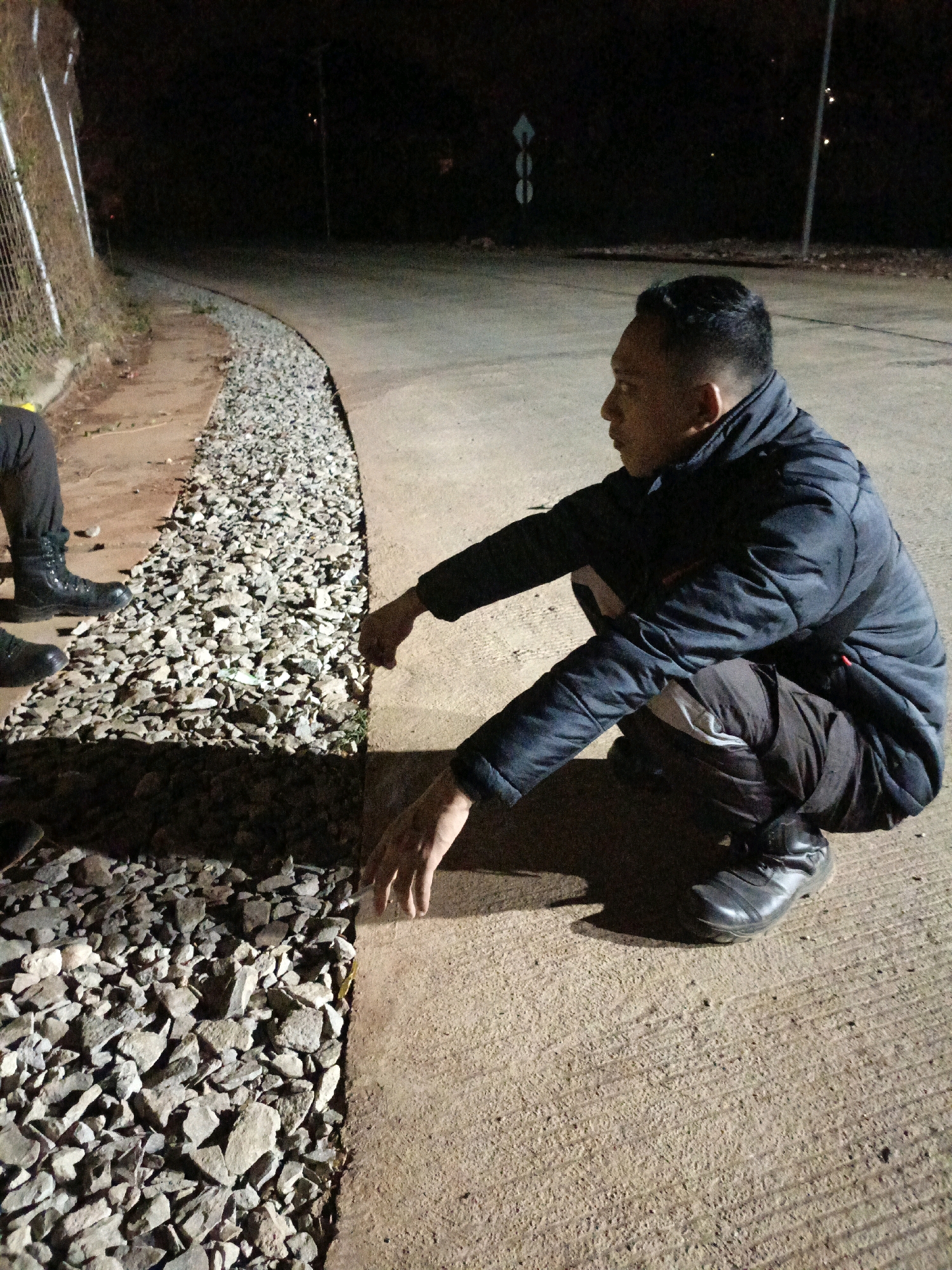 A man in a dark jacket and pants crouches on a paved surface next to a strip of gravel, illuminated by artificial light in Jenu, Indonesia. He is looking to his right, with his right hand reaching towards the gravel. In the background, a curved road is visible, with trees and darkness beyond. To the left of the gravel strip, another person's leg in dark pants and boots is visible, standing near a wire fence. The scene appears to be outdoors at night, possibly a roadside area.