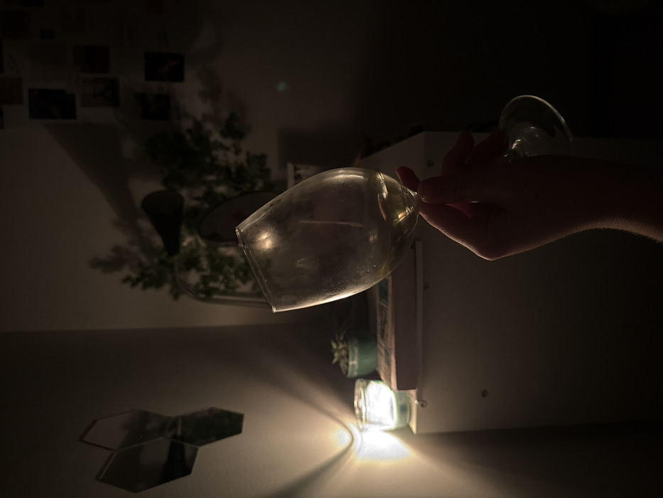 The image depicts an indoor scene in low light, identified as being within the Vozdovac Urban Municipality, Serbia. A human hand, positioned in the lower-center, holds a stemmed wine glass containing a small amount of pale liquid. The hand is partially silhouetted, with fingers wrapped around the glass stem and base.
The background shows a room corner. On the left, a light-colored wall features three dark, reflective, hexagonal wall decorations arranged geometrically. Below them, a lit candle in a clear glass jar illuminates a portion of a white or light-colored furniture surface, casting defined shadows on the wall. Adjacent to the candle on this surface is a small green succulent plant in a teal pot. A rectangular object, possibly a book or small box, rests behind the candle.
Further into the background, on the right, the wall is darker and displays several small, dark, rectangular items, possibly unframed photographs or prints. On a surface below these, likely an extension of the furniture from the left, are two larger potted plants, one appearing to be under a glass cloche. A dark, conical object is visible above one of these plants. The overall lighting is dim, primarily emanating from the candle, creating a focused illumination in the immediate vicinity of the candle and hand, with the rest of the room in shadow.