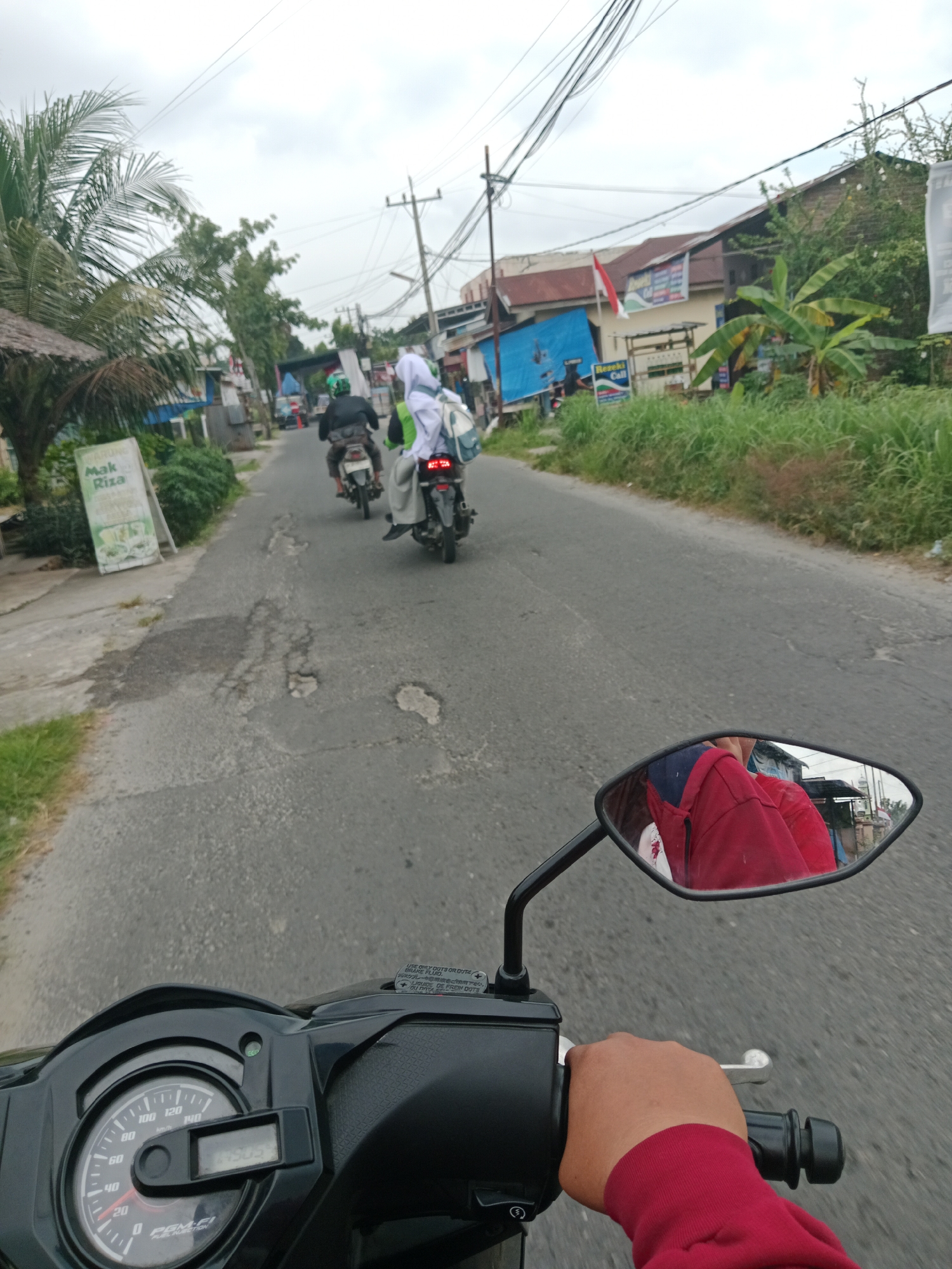 A first-person perspective shot captures a person riding a black scooter down a moderately damaged asphalt road in the City of Binjai, Indonesia. The sky is overcast, and there is lush green vegetation lining the right side of the road, with some buildings visible behind it. In the foreground, the scooter's handlebars and dashboard are visible, along with the rider's hand in a red long-sleeved shirt, gripping the throttle. A rear-view mirror reflects the rider's red top and part of their face, partially obscured by a dark blue head covering. Further down the road, two other scooters are visible, each carrying two passengers. The lead scooter has a rider in a black shirt and green helmet, while the second scooter has a passenger in a white headscarf and matching white garment. A sign with "Mak Riza" is visible on the left side of the road. The road surface is rough with several potholes and cracks. Power lines are strung overhead.