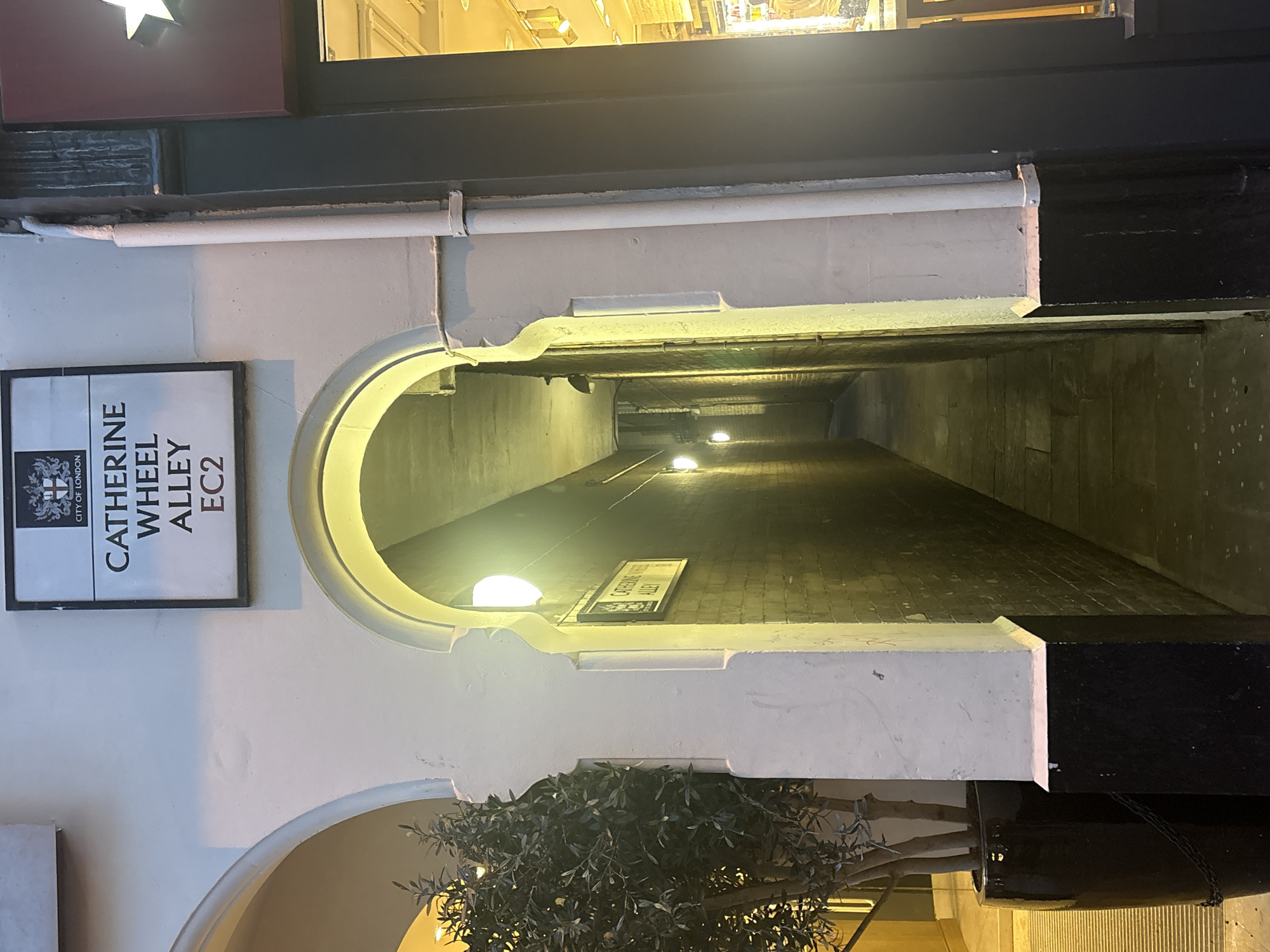 The image depicts Catherine Wheel Alley, EC2, in the City of London. The alleyway is viewed through an arched entrance, with a white frame and black trim. The entrance features a sign on the left side indicating the alley's name and postcode, "CATHERINE WHEEL ALLEY EC2," as well as an official logo for the City of London. A plant is situated below the arch. The alleyway is long and narrow, dimly lit by lights placed along its length. The walls appear to be made of brick or stone, and there is a sign bearing the street name visible further down the alleyway.