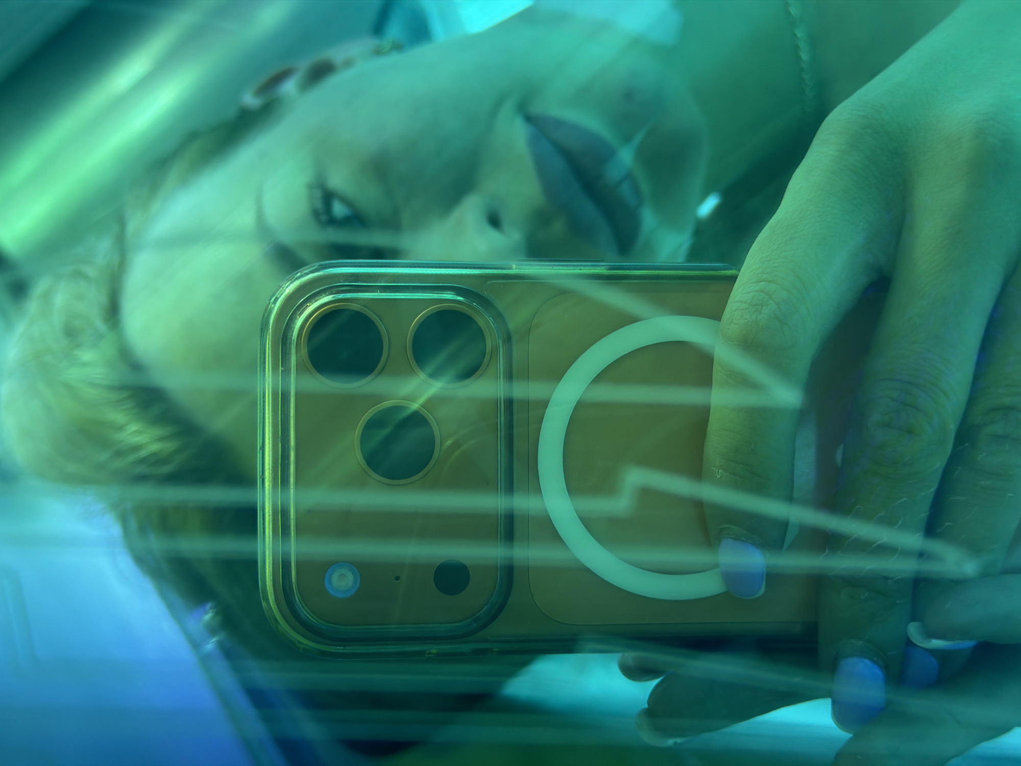 A person is tanning in a tanning bed, taking a selfie with their phone. The tanning bed has green fluorescent lights. The person is lying down and looking at their phone, which is held in their hand. The phone has a camera system with multiple lenses and a magnetic ring. The person's face is visible in the reflection of the tanning bed's acrylic cover, appearing blurred due to the lighting and angle. The person's hand is also visible, holding the phone and showing painted fingernails. The environment is enclosed and illuminated by artificial light, suggesting an indoor tanning salon. The image does not provide any specific information about the location being in the city of Kyiv, Ukraine.