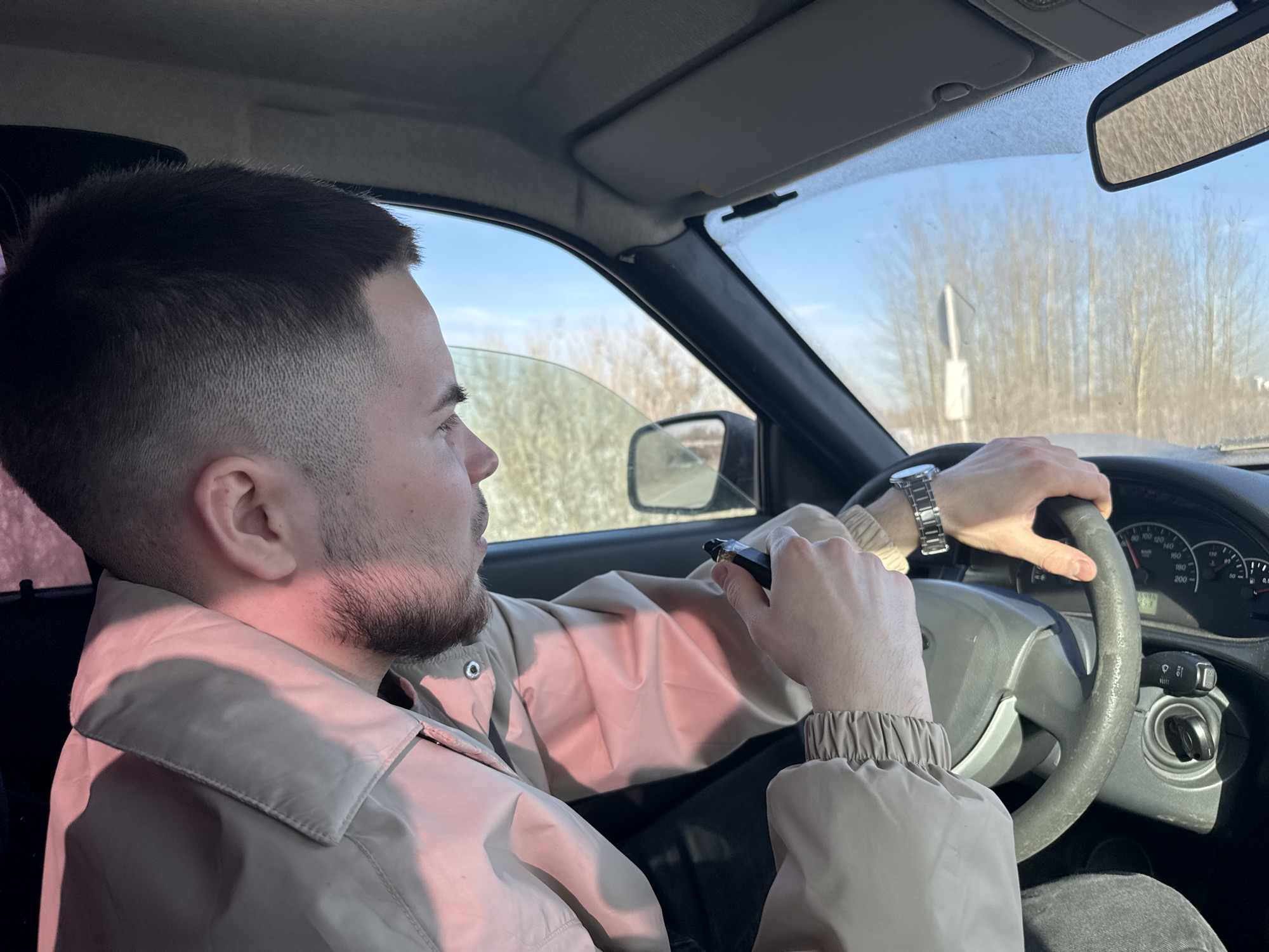The image depicts a male individual seated in the driver's seat of a vehicle in Кондратово, Russia. The man, seen from a side profile facing right, has short dark hair with a fade haircut and a short beard. He is wearing a light tan or beige collared jacket, which shows a distinct pinkish-red hue from sunlight entering the vehicle. A silver-toned wristwatch is visible on his left wrist. His left hand rests on the top portion of the vehicle's dark gray steering wheel, while his right hand holds a small black electronic device, possibly a vape pen. The instrument cluster is visible, indicating a speedometer ranging up to 200 km/h, a tachometer, and other gauges, with the speedometer needle resting at zero. The windshield is noticeably dusty, through which a clear blue sky is visible above a sparse stand of bare deciduous trees. A white rectangular road sign with an angled top is partially visible in the distance to the left of the trees. The vehicle's interior features a light gray fabric headliner, a matching sun visor, a black A-pillar, and a rear-view mirror. The scene is illuminated by natural daylight, with strong sunlight casting a warm glow from the side.