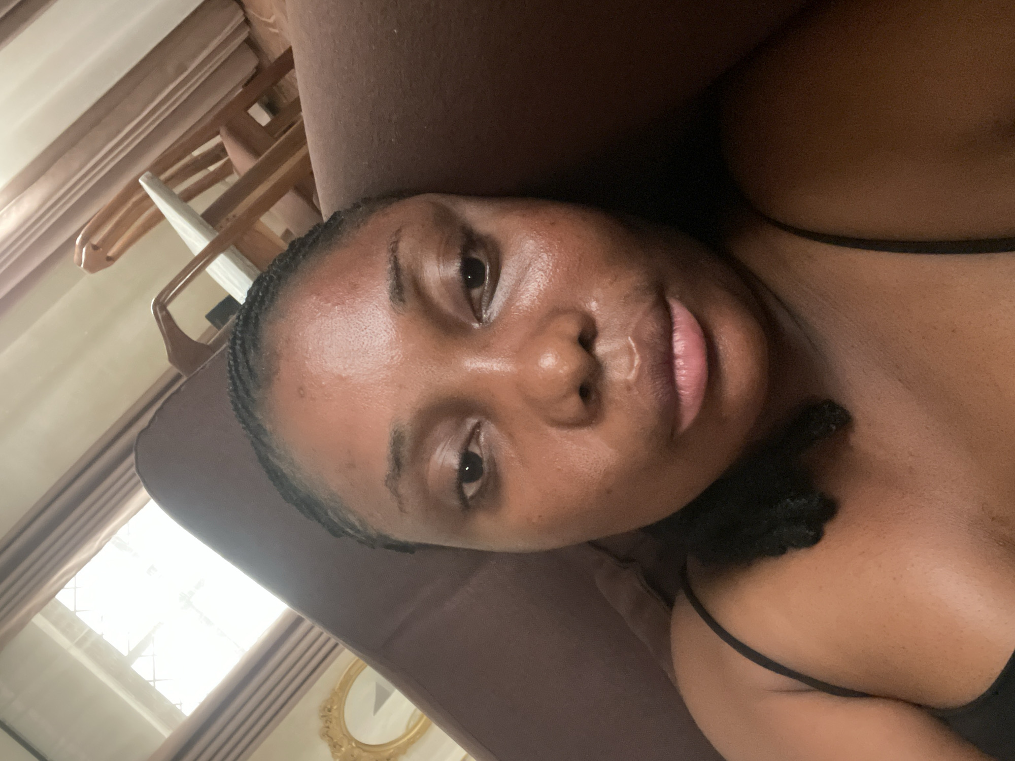A medium close-up, portrait-oriented photo shows a young woman with dark skin taking a selfie while lying down or leaning back. Her head is tilted to the left in the frame. She has braided black hair, pulled back from her face. Her eyes are open and dark, looking directly at the camera. Her lips are full, slightly parted, revealing a natural pinkish-red hue. Her skin appears moisturized, with some natural shine on her forehead, nose, and cheeks. She is wearing a black top with a thin strap visible over her right shoulder and chest area, which is partially shown in the lower right of the frame.
The background is softly blurred but reveals details of an indoor setting. To the upper left, bright daylight streams through a window with a grid pattern, indicating it is daytime. Light-colored curtains frame the window. Below the window, wooden structures resembling hangers or parts of furniture are discernible. To the lower right, a portion of an ornate, gold-framed mirror is visible. The surface she is resting on appears to be a dark brown, possibly fabric, like a sofa or bed. The overall impression is a relaxed, casual moment indoors, likely in a home in Jos, Nigeria. No visible text is present in the image.