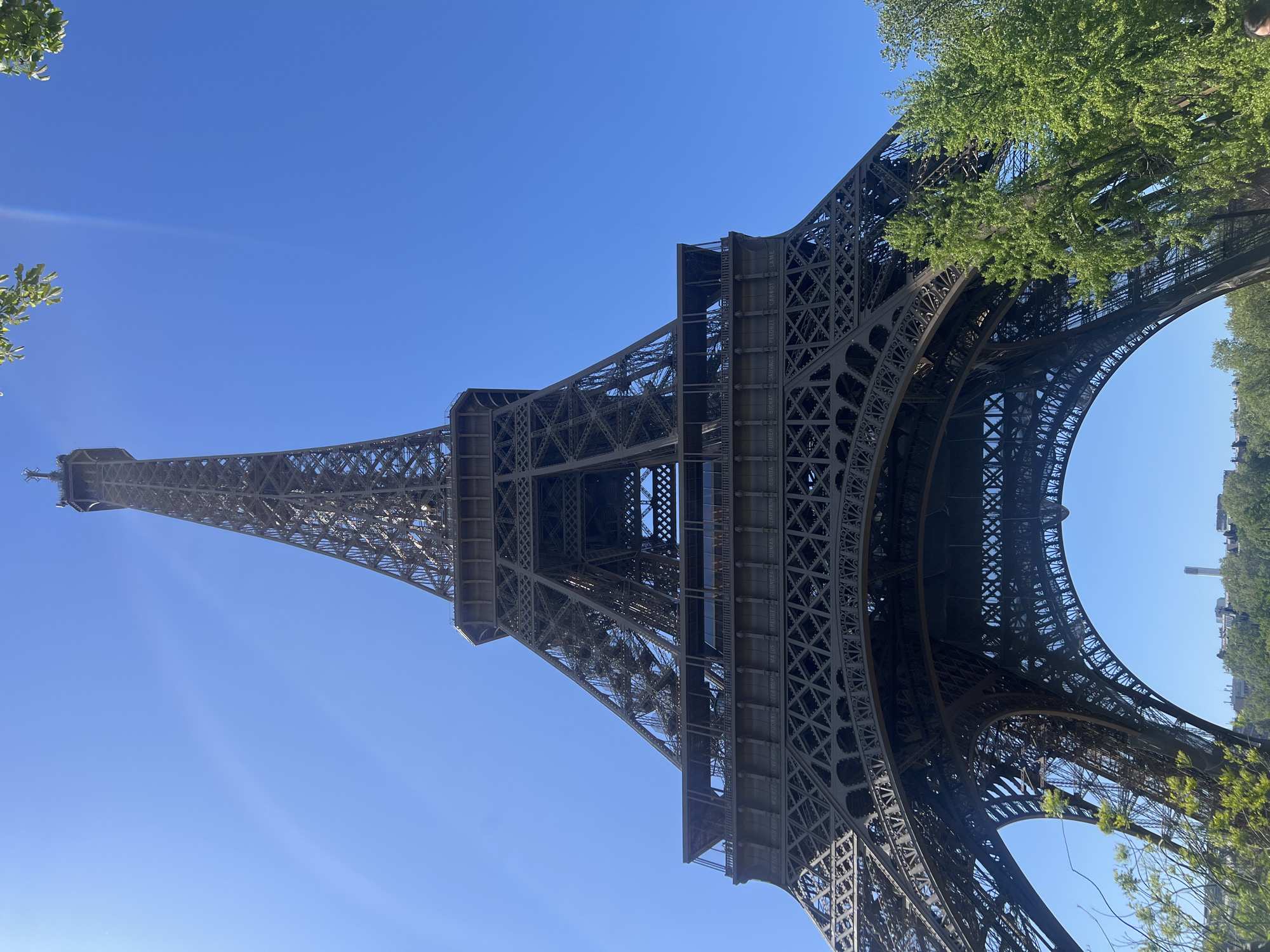 This media file captures a low-angle view of the Eiffel Tower in Paris, France, against a clear, vibrant blue sky. The image is taken from directly beneath or very close to the base of the tower, emphasizing its immense height and intricate iron lattice structure.
The primary subject is the Eiffel Tower, with its dark, complex metal framework dominating the composition. The photograph encompasses the lower sections of the tower, including its distinct arches and the first and second platforms, extending upwards towards its narrow spire. Green foliage from trees is visible on the left and right sides of the frame, partially obscuring the tower's base and framing the sky, suggesting a park-like or tree-lined setting around the monument.
The scene depicts a bright, sunny daytime, indicated by the brilliant blue sky and the clear, crisp lighting on the tower's structure. There are no clouds visible, and subtle lens flares or sunbeams can be seen in the sky, suggesting strong sunlight. No people or specific activities are clearly depicted, placing the sole focus on the architectural landmark.
A notable detail is the presence of engraved names on the horizontal girders of the tower's first level. These are the names of 72 French scientists and engineers. Clearly legible names include "CARNOT," "FOURIER," "BIGAR," "PELOUZE," "SAUVAGE," "CLAPEYRON," "LAME," "SAINT-VENANT," "MASSON," "BRÉGUET," "DUGAS," "MALUS," and "DE LA GRANDE." The photograph, which appears to be rotated 90 degrees clockwise, offers an up-close and immersive perspective of this iconic Parisian landmark.