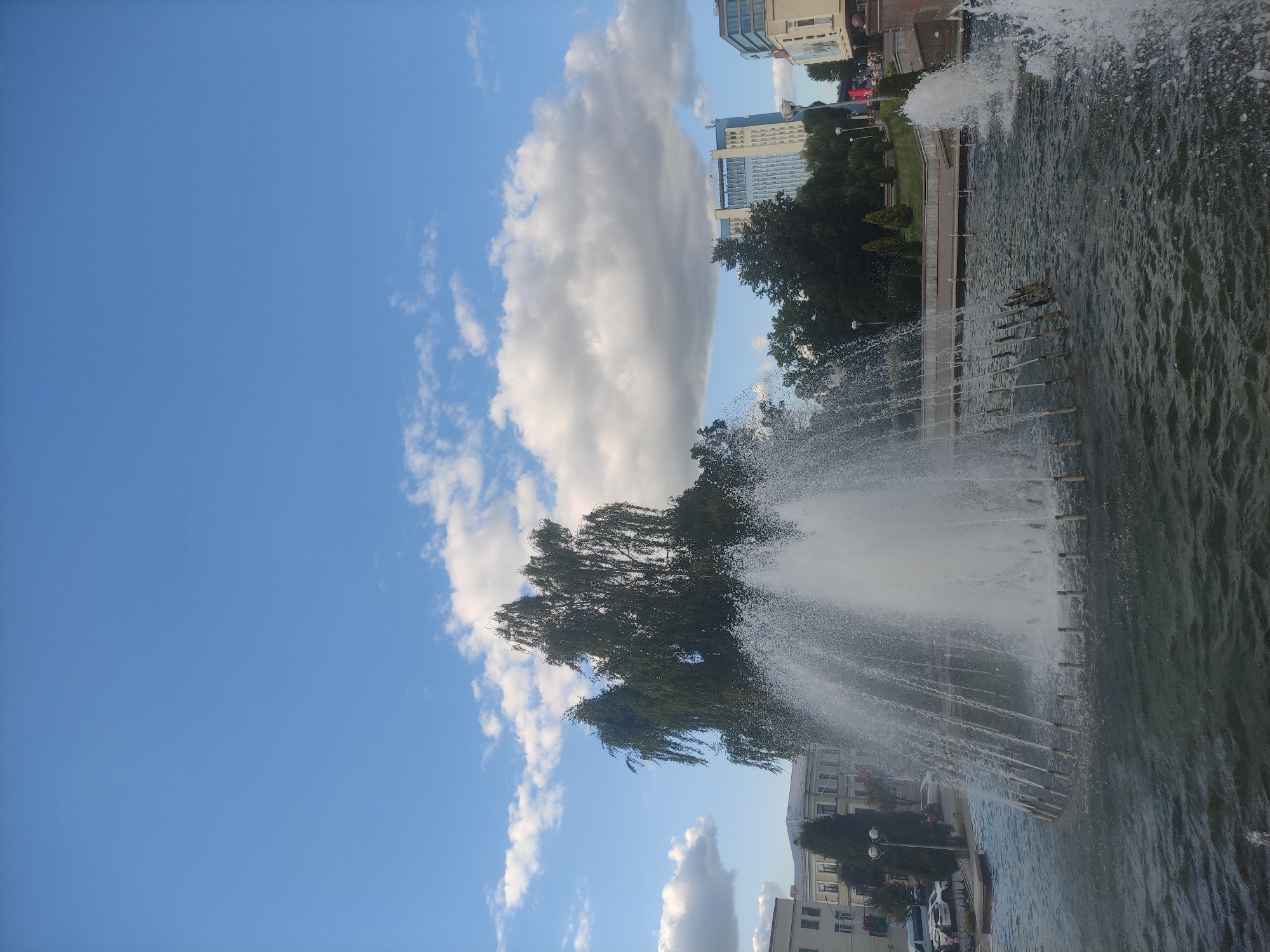 This is a medium-distance, eye-level shot of a large public fountain in Mahilyow, Belarus, with a bright blue sky filled with fluffy white clouds above. The fountain is the dominant subject, with water spraying upwards in a large, multi-tiered arc, creating a misty effect. Several smaller water jets are also visible in the foreground and to the right. The fountain is situated in a body of dark, rippling water. Behind the fountain, a large, leafy tree with drooping branches stands prominently. In the background, several buildings are visible. To the left, a light-colored, multi-story building with visible windows is partially obscured by the tree. Further back and to the right, a tall, modern blue and white building with many windows stands out, likely an office building or hotel. Smaller trees and manicured green spaces are also present in the mid-ground. Streetlights are visible near the buildings. A few cars are parked on a road to the far left. The overall scene depicts a sunny day in an urban park or public square, likely a popular spot for recreation.