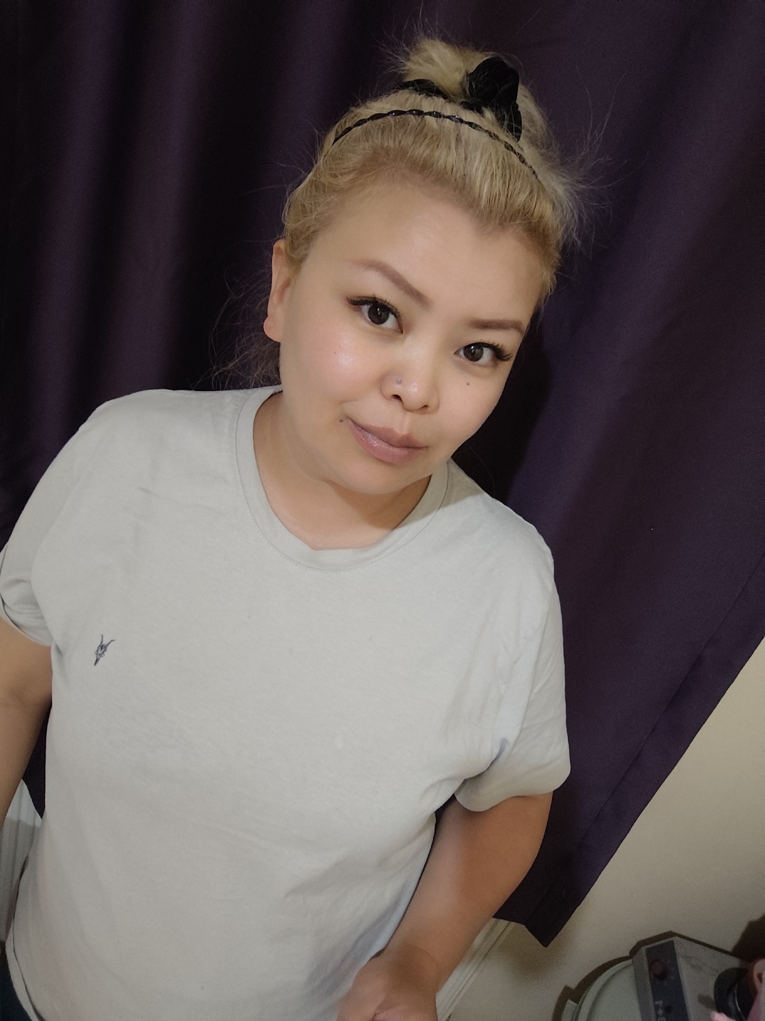 A woman is pictured indoors, facing the camera. She has blonde hair pulled back with a headband and a black bow. Her t-shirt is light-colored, and a dark-colored curtain is visible in the background. A small logo is visible on the t-shirt. The location is Unknown, Unknown.