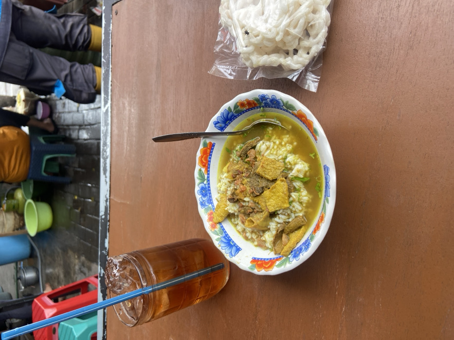 A bowl of Indonesian soto, a traditional soup with rice, meat, and vegetables, sits on a brown surface alongside a glass of iced tea with a blue straw. A package of white crackers is placed at the top of the frame. The table is outdoors, with a glimpse of a bustling street scene including seating, equipment, and pedestrians in the blurred background.