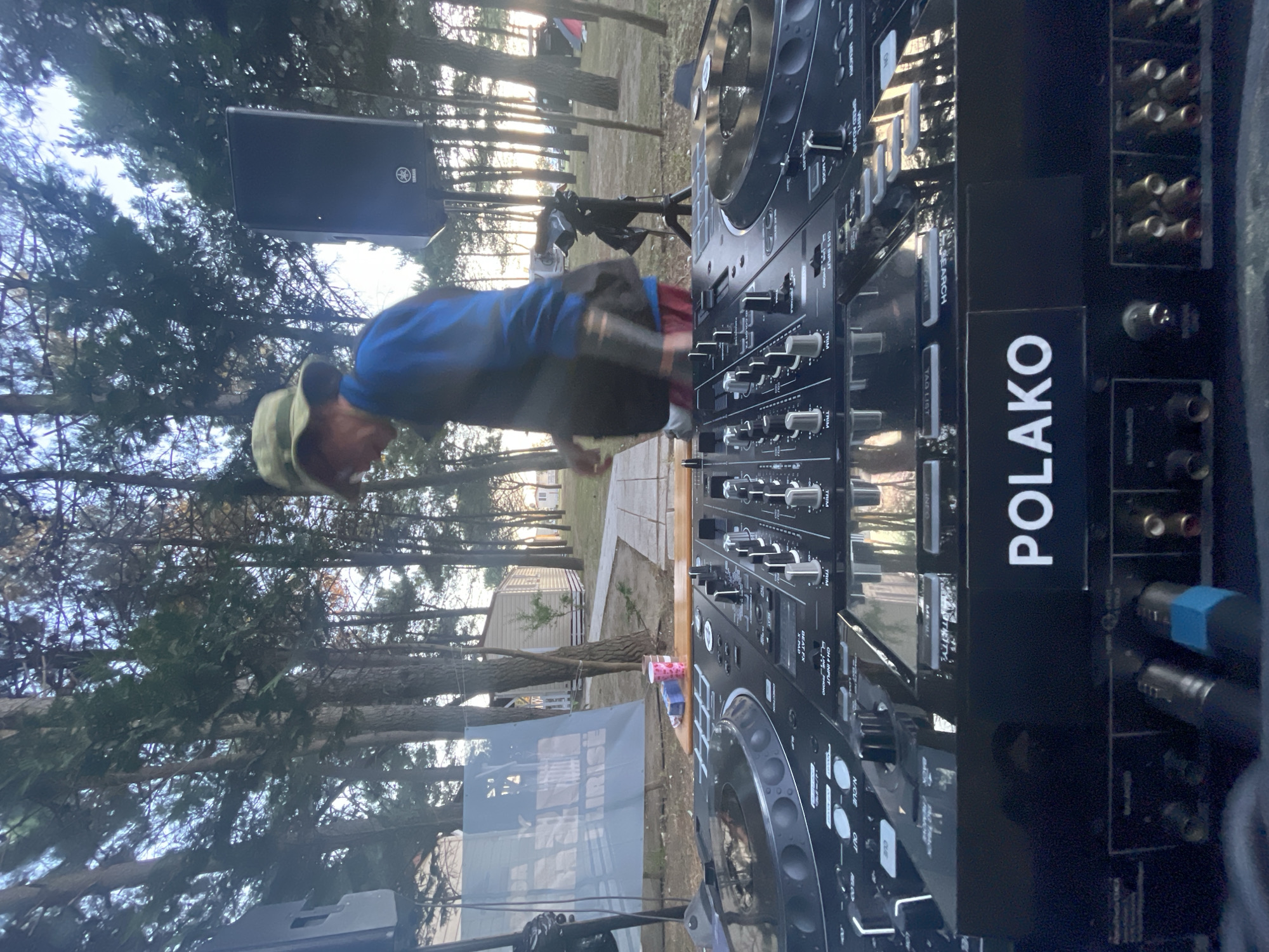 The image shows a person standing in front of a DJ setup in an outdoor location. The DJ setup includes a mixer with the name "POLAKO" on the side. A speaker is mounted on a stand behind the person. The environment appears to be a park or wooded area with trees and a building in the background.