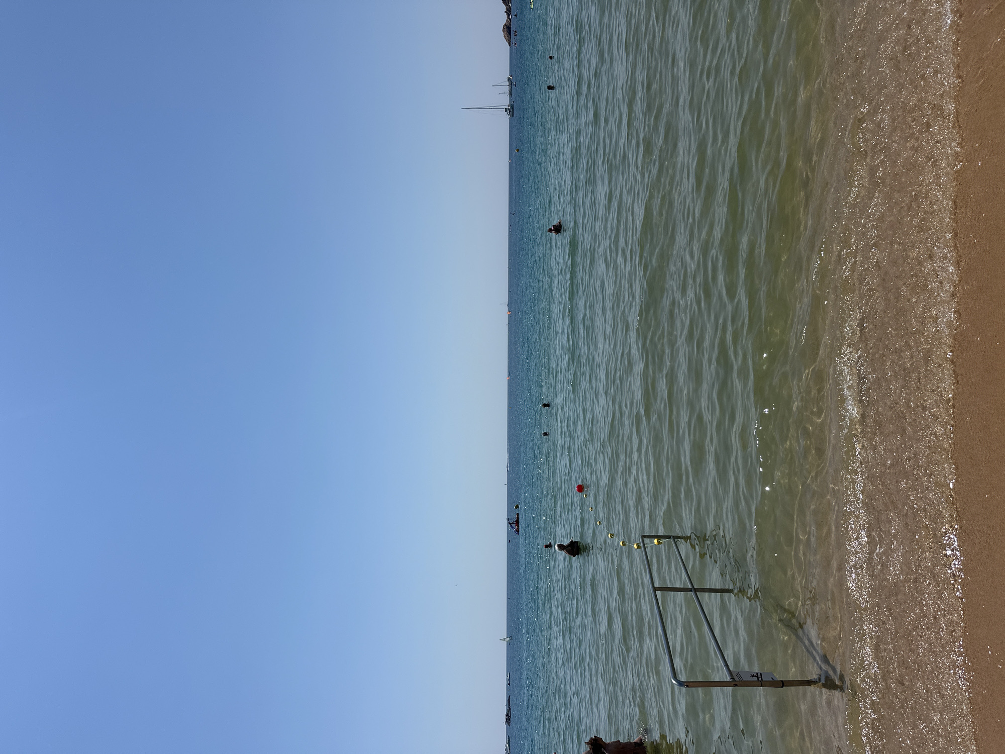 The image depicts a serene beach scene in Sant Feliu de Guíxols, Spain, under a clear blue sky. In the foreground, a metal railing, partially submerged in the shallow, clear water, extends from the sandy shore into the sea. Small yellow buoys on a rope demarcate a swimming area.
Several individuals are visible in the water, some swimming, others standing. In the middle ground, a person is visible with their head and shoulders above the water, facing away from the camera. Further out in the sea, several boats are present, including a sailboat, a motorboat, and what appears to be a jet ski. The distant horizon is marked by a line of islands or coastline. The water is calm and appears to be a tranquil turquoise color, reflecting the bright sunlight. The sand is a warm, golden brown, with small waves gently lapping at the shore. The overall impression is one of a peaceful and enjoyable day at the beach.