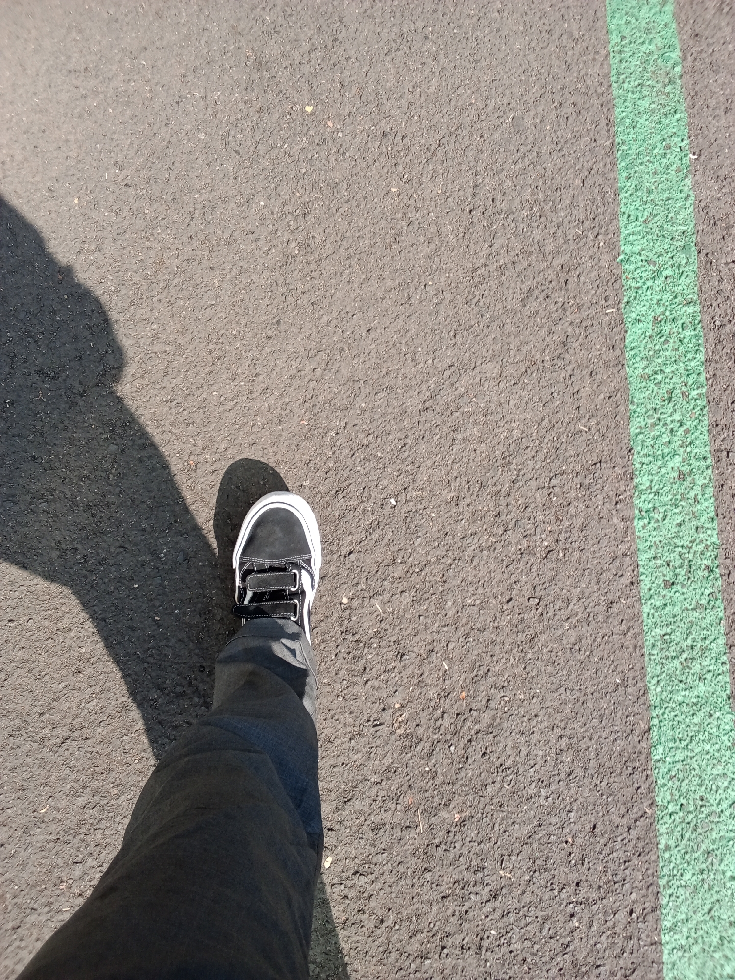 The image shows a close-up, high-angle view of a person's left leg and foot wearing a black and white sneaker and dark pants, standing on an asphalt surface. A bright green line is painted on the asphalt to the right of the foot. A shadow of a person is cast on the asphalt to the left. The scene appears to be outdoors, likely on a paved area such as a sidewalk, road, or sports court. The asphalt is textured and gray, with some small pebbles visible. There is a subtle shadow from the person's foot. No specific landmarks or indicators are present to pinpoint the exact location within Bandung City, Indonesia.