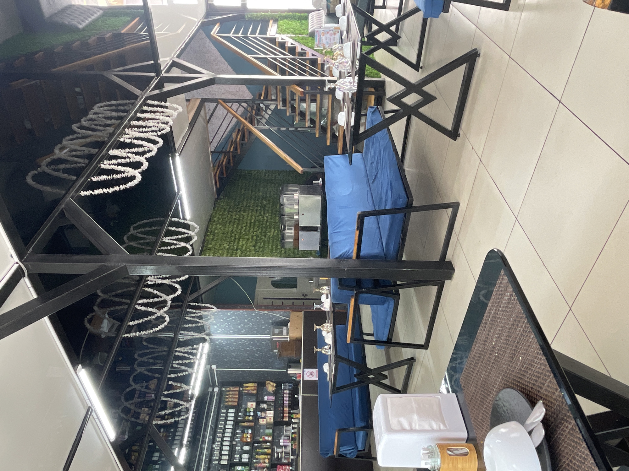 The image depicts the interior of a restaurant located in Bosteri, Kyrgyzstan. The dining area features blue upholstered sofas and chairs arranged around black metal-framed tables with glass tops. Several tables are set with white plates and glassware. In the background, a bar area is visible with shelves stocked with bottles. A set of wooden stairs with black railings leads to an upper level. The walls are decorated with textured green panels. Overhead, decorative white garlands are hung. The floor is tiled in a light color. The scene is brightly lit by artificial lighting.