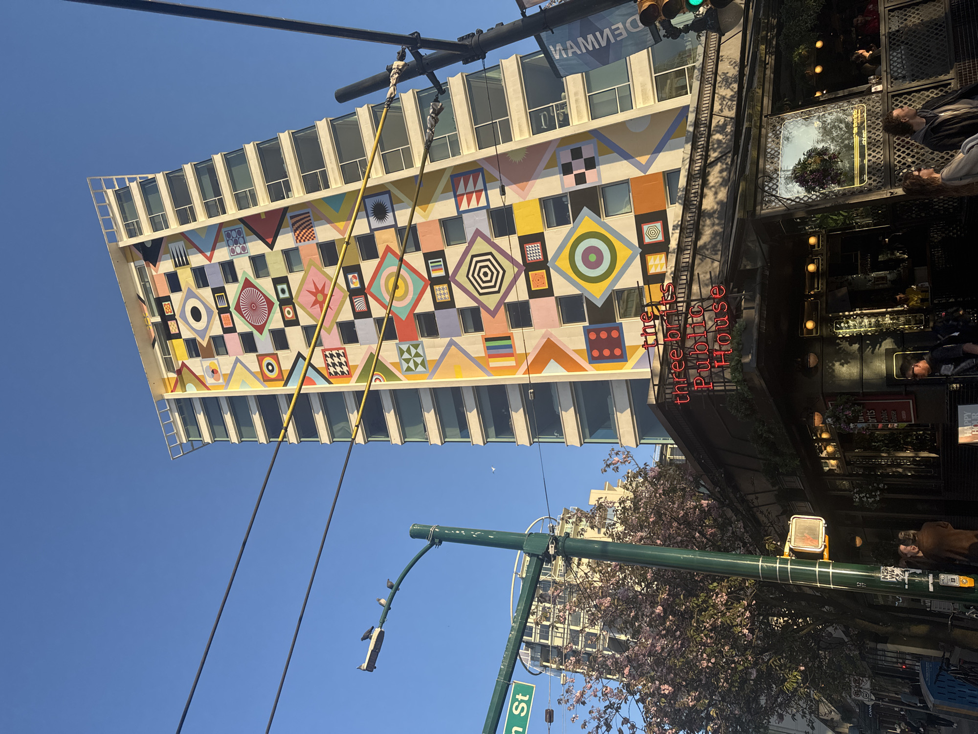 The image shows a building with a colorful geometric mural in Vancouver, Canada. The mural covers a large section of the building's facade, contrasting with the surrounding architecture. Street signs reading "Hamilton St" are visible. The text on the mural reads "DENMAN". A neon sign reads "the three brits Public House". Pedestrians are visible in the image.