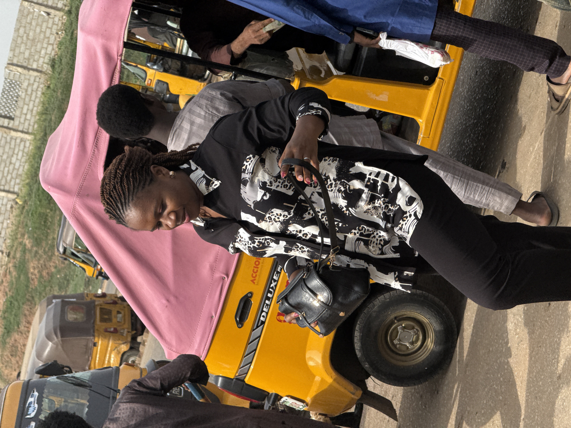 The image, taken in Abuja, Nigeria, captures an active outdoor scene centered around a vibrant yellow commercial tricycle (keke napep) with a pink canvas canopy. The main subject is a woman standing beside the tricycle, near its rear wheel. She has braided hair and is dressed in a black long-sleeved top featuring a distinct white and black abstract or animal print pattern, paired with black trousers. She holds a black handbag by its strap.
Several other individuals are visible around and inside the tricycle. One person inside wears a blue top and appears to be holding money or papers. Another individual with short, dark hair is seen near the vehicle, wearing a light-colored striped top. The legs and feet of multiple people, some wearing sandals or flip-flops, are also visible on the asphalt ground.
The side of the yellow tricycle clearly displays the words "ACCIO" and "DELUXE" printed vertically. In the background, other yellow tricycles are present, suggesting a busy street or a transport hub. The environment includes a rough concrete block wall and some sparse vegetation in the distance. The scene is brightly illuminated by natural daylight, casting clear shadows on the ground, indicating a daytime setting. The image portrays a slice of daily urban life, likely at a public transport stop.