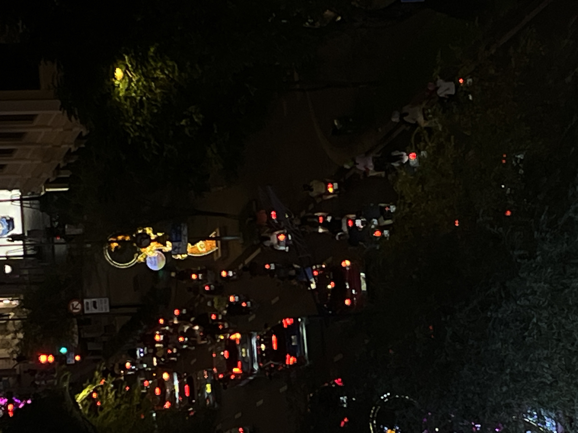 This is a high-angle, nighttime aerial view of a busy street in Thủ Đức, Vietnam, filled with motorbikes and cars. The street is illuminated by headlights, taillights, and the colorful lights of a decorative structure. Trees line the side of the road, their leaves appearing green and yellow in the artificial light. The scene depicts a bustling urban environment with vehicles moving in both directions.