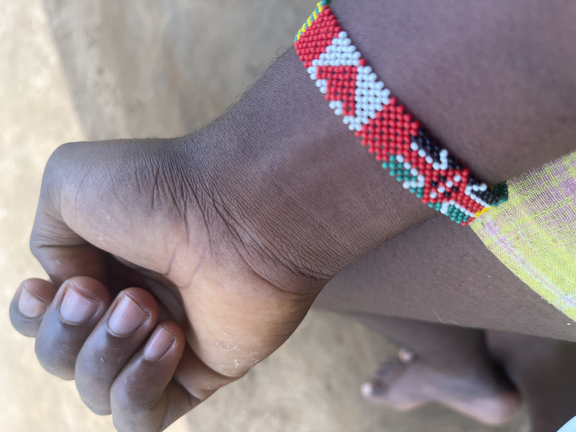 The image shows a person's dark-skinned arm with a tightly clenched fist. A colorful, beaded bracelet adorns the wrist, featuring patterns in red, white, black, and green, bordered by yellow beads. A piece of yellow fabric is visible adjacent to the bracelet. Part of the person's foot is also visible in the background. There is no information to determine the location.