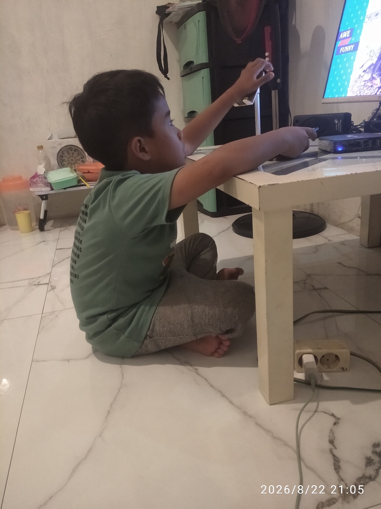 A young boy with dark hair sits on a white marble floor, facing a white table and a computer screen. He is wearing a light green t-shirt and grey pants. His left hand holds a silver smartphone, while his right hand rests on the table, pointing towards a black device. The computer screen displays a video with the words "AWE" and "FUNNY" visible. A white electrical outlet with a plugged-in grey cable is located near the base of the table. The background shows a green storage unit and a dark speaker. The scene is set indoors, likely in a residential area, and the timestamp "2026/8/22 21:05" is present in the lower right corner, suggesting it was captured in Nglebak, Indonesia.