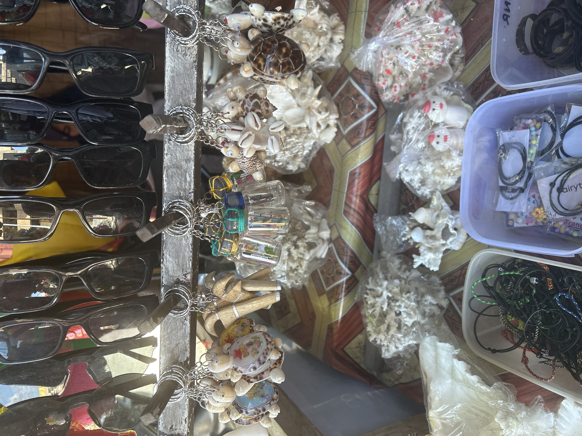This is a close-up, eye-level shot of a street stall in Bira, Indonesia, displaying various souvenirs. The stall features a row of sunglasses hanging from hooks at the top. Below the sunglasses, a variety of keychains are displayed on a wooden bar. These keychains include small turtle figurines made of shells, wooden totems, and tiny glass bottles filled with what appear to be sand or shells. The background reveals more merchandise, including decorative shells, corals, and various bracelets or necklaces in plastic containers. The overall impression is one of a tourist-oriented market stall.