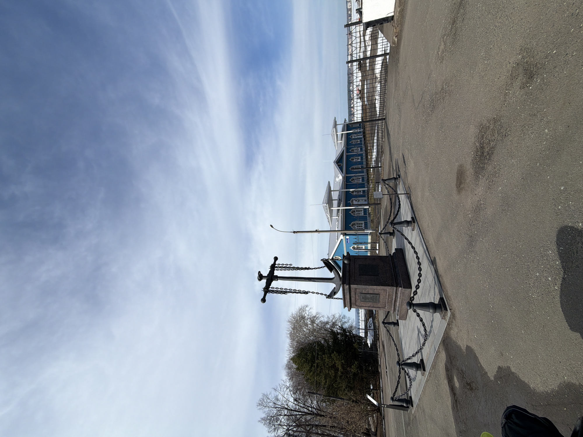The image shows a monument consisting of a large anchor mounted on a stone pedestal, surrounded by a chain fence on a paved surface. The monument is located in the city of Votkinsk, Russia. Behind the monument are leafless trees and a blue wooden building with white trim. A metal fence is visible beyond the building. The sky is partially cloudy with a mix of blue sky and wispy clouds. Part of the photographer's body is visible in the bottom right corner of the image.