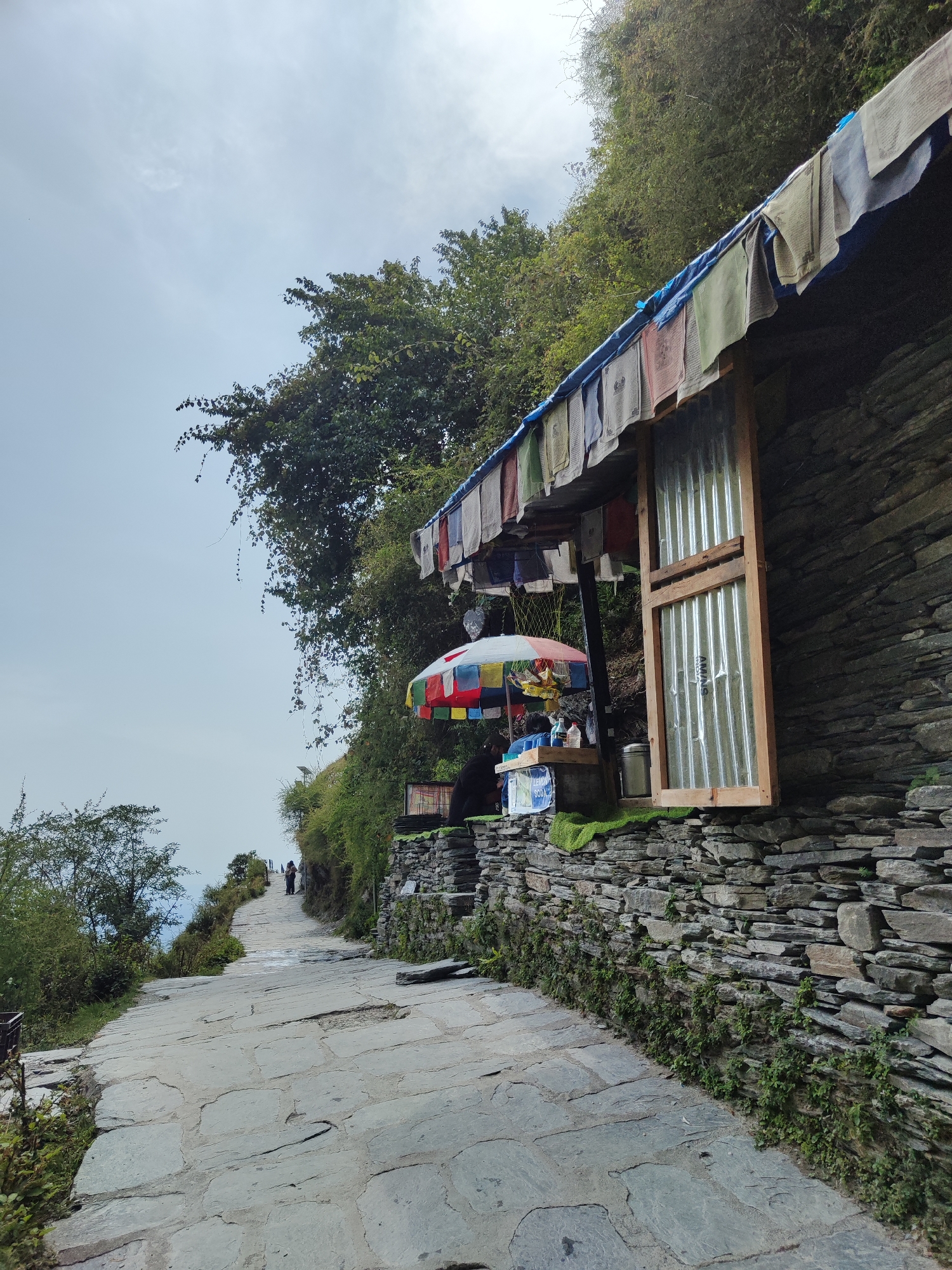 The image captures a stone-paved pathway winding uphill through a hilly or mountainous landscape. On the right side of the path, a sturdy building constructed from stacked flat stones is nestled against the hillside. Its roof is covered by a blue tarp and adorned with a long string of colorful Buddhist prayer flags.
Underneath the roof, a small stall or shop is visible, featuring a vibrant, multi-colored umbrella (red, blue, yellow, green, and white sections) providing shade. Behind the counter, a person is partially obscured. On the stall, various bottles and a metal container are present. A small white sign clearly displays the text "LEMON 500," likely indicating the price for lemonade. Adjacent to the stall is a window or door opening with a wooden frame enclosing a corrugated metal panel, on which the letters "AMNS" are visible.
The path itself is irregularly paved with large, grey, flat stones and ascends gradually. Lush green trees, bushes, and smaller plants line both sides of the pathway and densely cover the steep hillside behind the building. In the distance, further up the path, two small figures can be seen walking, suggesting a trekking trail or a route connecting mountain settlements. The sky is largely overcast and hazy, casting a soft, diffused light over the scene, indicating daytime, possibly late morning or early afternoon. The presence of prayer flags strongly suggests a location within a Buddhist cultural region, such as the Himalayas.