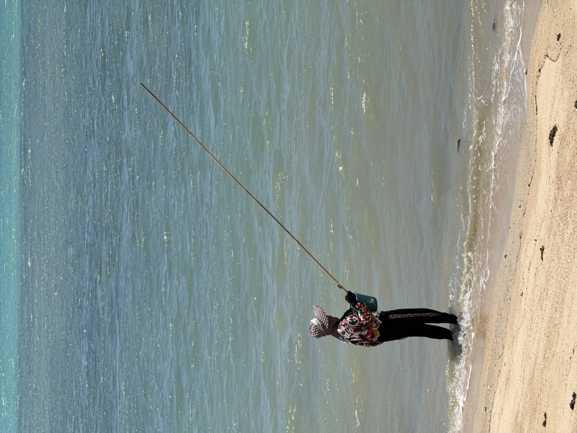 A person stands in the shallow water at the edge of a sandy beach, fishing with a long pole. The person is wearing a checkered hat, a brightly colored floral shirt, dark pants, and gloves. They are holding a blue basket. The water is choppy and a light turquoise, with whitecaps indicating a windy day. The beach is light brown sand with a few pieces of dark debris scattered on it. The background shows a gradient of turquoise water, becoming a deeper blue further out. The scene is likely on the coast of Ko Samui District, Thailand, given the typical weather and environment.