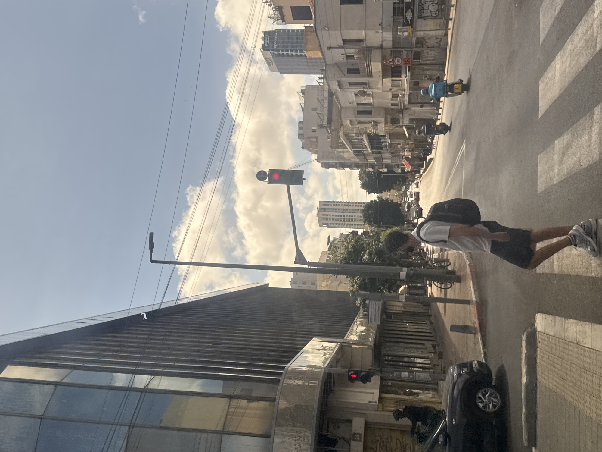The image depicts an urban scene in Tel Aviv, Israel. A pedestrian with a backpack, wearing a white shirt and dark shorts, is walking across a crosswalk. In the background, a street extends into the distance, lined with buildings of varying heights. A traffic light shows a red signal. A dark car is parked on the side of the road. The sky is visible, with a mix of blue and clouds.