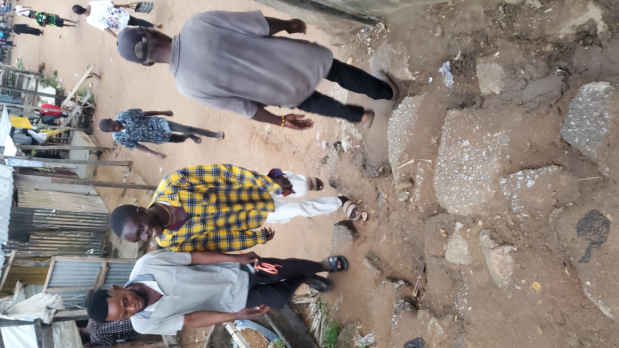 The image captures a street scene in Jalingo, Nigeria, likely in a commercial or residential area characterized by informal structures and uneven terrain. In the foreground, three young men are walking uphill on a path of dirt and broken concrete. The man on the left, wearing a light grey polo shirt and black pants, is looking down and appears to be smiling. Next to him, a man in a yellow and black plaid shirt and white pants is also looking down and smiling. The third man, in the rear, is wearing a dark blue patterned shirt and dark pants.
The environment consists of a narrow path with scattered rocks and debris. To the left, there are corrugated metal structures and makeshift stalls, suggesting a market or informal settlement. In the background, other people are visible, some walking and some standing, and more structures with corrugated metal roofs are present. The overall impression is one of a busy, somewhat disorganized urban environment.