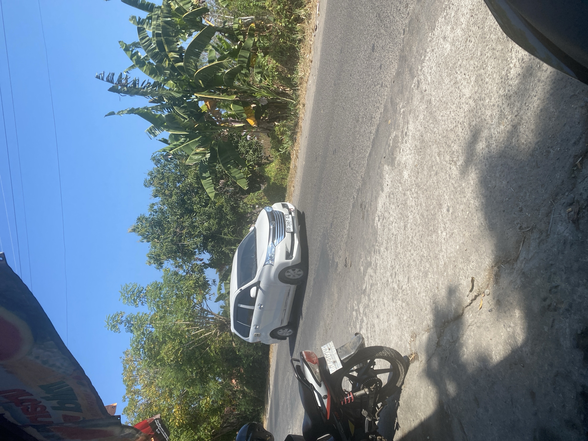 A white sedan is driving down a paved road in Mattekko, Indonesia, with lush green trees and banana plants lining the sides of the road. The sky is clear and blue. In the foreground, a motorcycle is parked on the side of the road. The overall scene is bright and sunny.