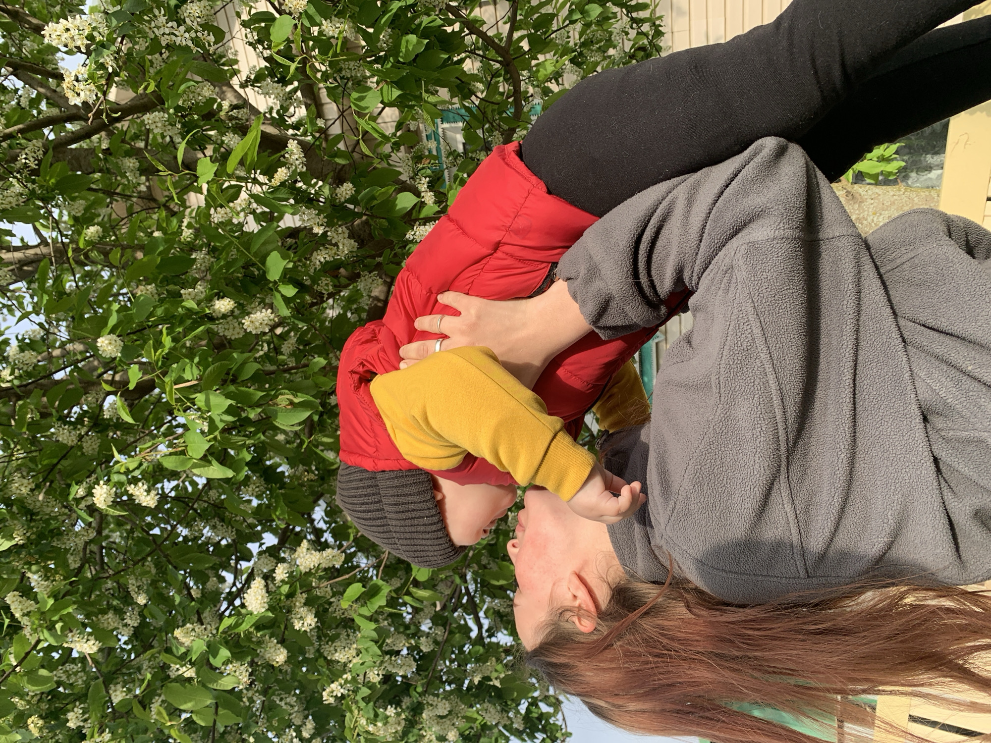 A woman with red hair wears a gray fleece jacket while holding a child who is wearing a black knit hat, a red puffer jacket, and a yellow sweatshirt. The pair is positioned in front of a blossoming tree with white flowers. The scene appears to be outdoors, with a building visible in the background. The exact location in Фоки, Russia, cannot be determined from the image. The woman and child are likely interacting in a familial manner.