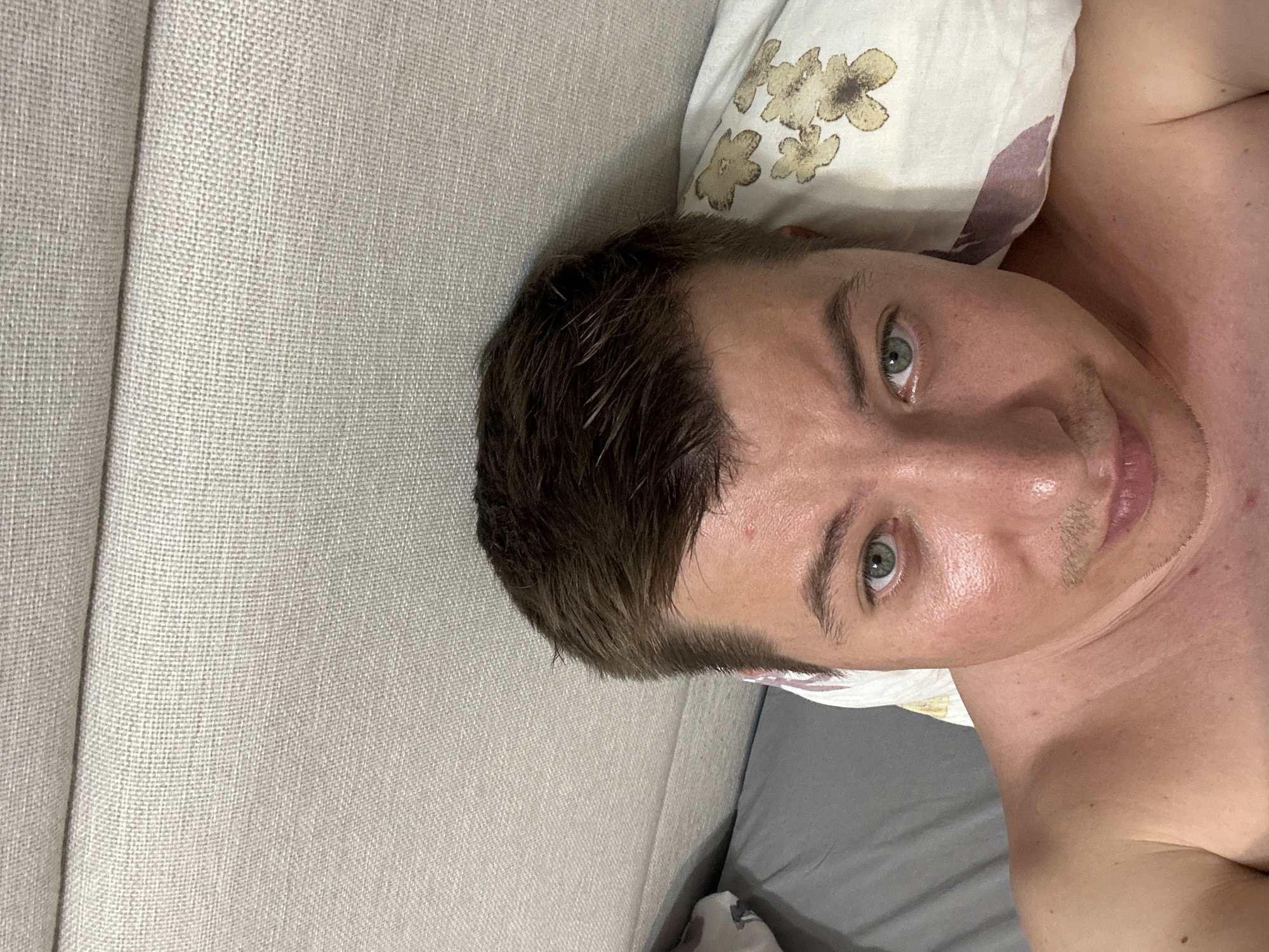 The image shows a close-up, high-angle selfie of a man lying on a bed, possibly in Budva, Montenegro. He has short, dark brown hair and green eyes, with a light complexion and a faint mustache. He is shirtless, and his right shoulder and chest are visible. The man is positioned with his head resting on a pillow that has a floral pattern in muted tones. Behind him, the bed is covered with a textured, light-colored fabric, likely a headboard or wall upholstery. To the left of the man, a gray pillow or duvet is partially visible. The overall setting suggests an indoor environment, likely a bedroom.
