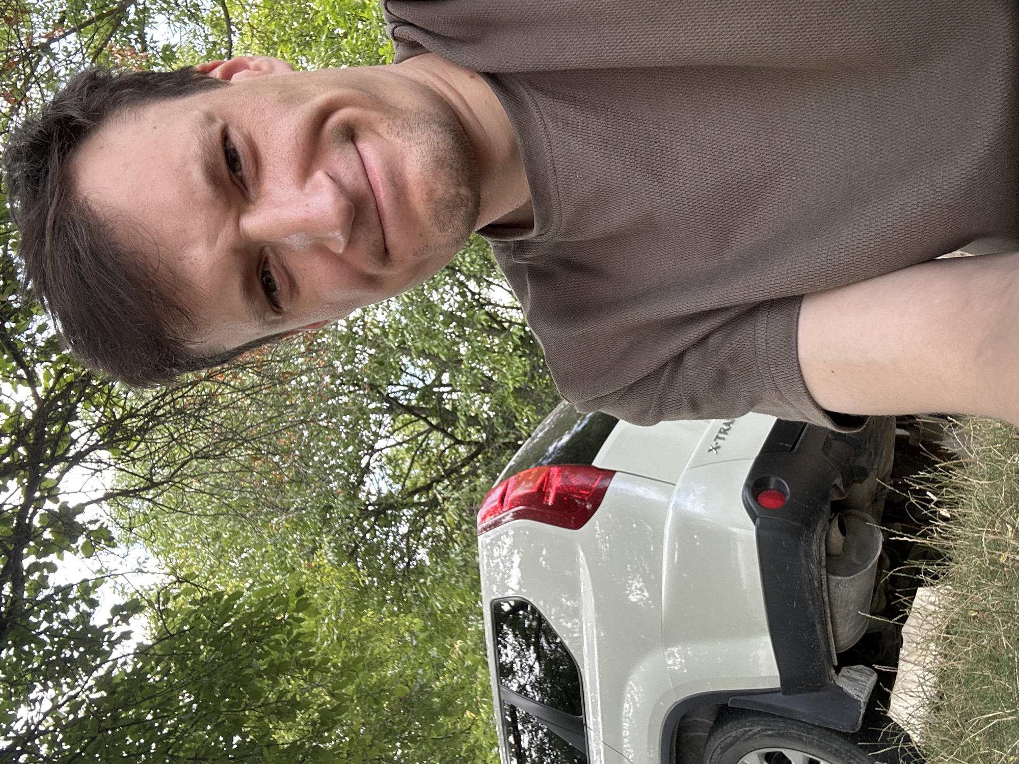A man with short brown hair and a slight stubble is taking a selfie outdoors. He is wearing a brown, short-sleeved t-shirt with a textured pattern. The man's left side is partially obscured by the back of a white SUV. The SUV has a red taillight and the lettering "X-TRAIL" visible on the rear bumper. The background consists of lush green trees and foliage, suggesting a rural or park-like setting. The ground beneath the SUV and in the foreground is covered with dry grass and some rocks. The lighting indicates it is daytime, possibly overcast or in a shaded area due to the leaves overhead. The man's expression is a slight grimace.