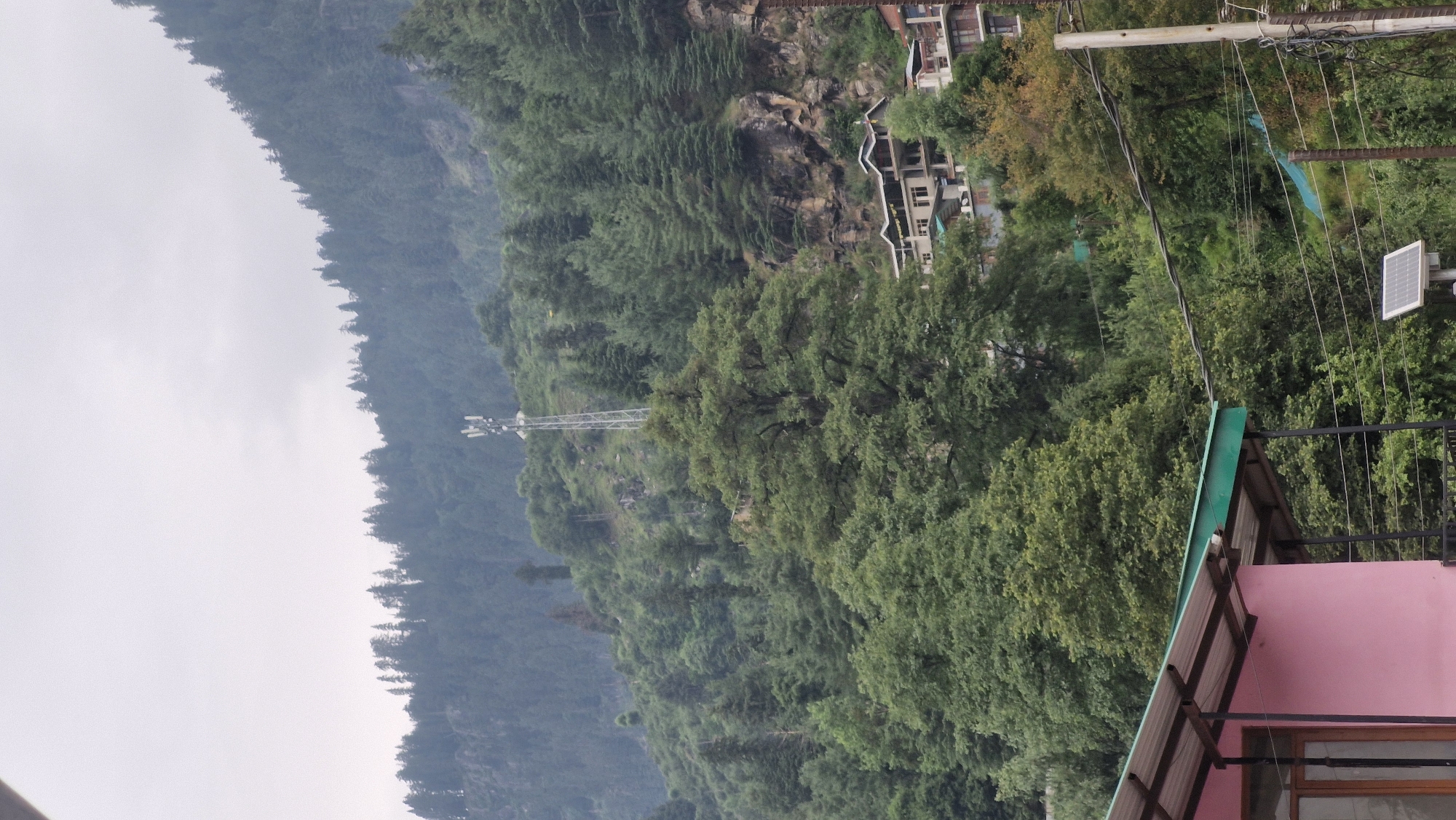 The image captures a view of a hillside in Vashisht, India, with lush green trees covering the slopes. In the middle ground, a cell tower stands tall, indicating a populated or developing area. Several buildings with multi-story structures and slanted roofs are visible on the hillside, suggesting a settlement integrated with the natural landscape. In the foreground, a portion of a pink building with a green-roofed overhang is visible, and a utility pole with electrical wires and a small solar panel are present. The sky is overcast, casting a diffused light on the scene.